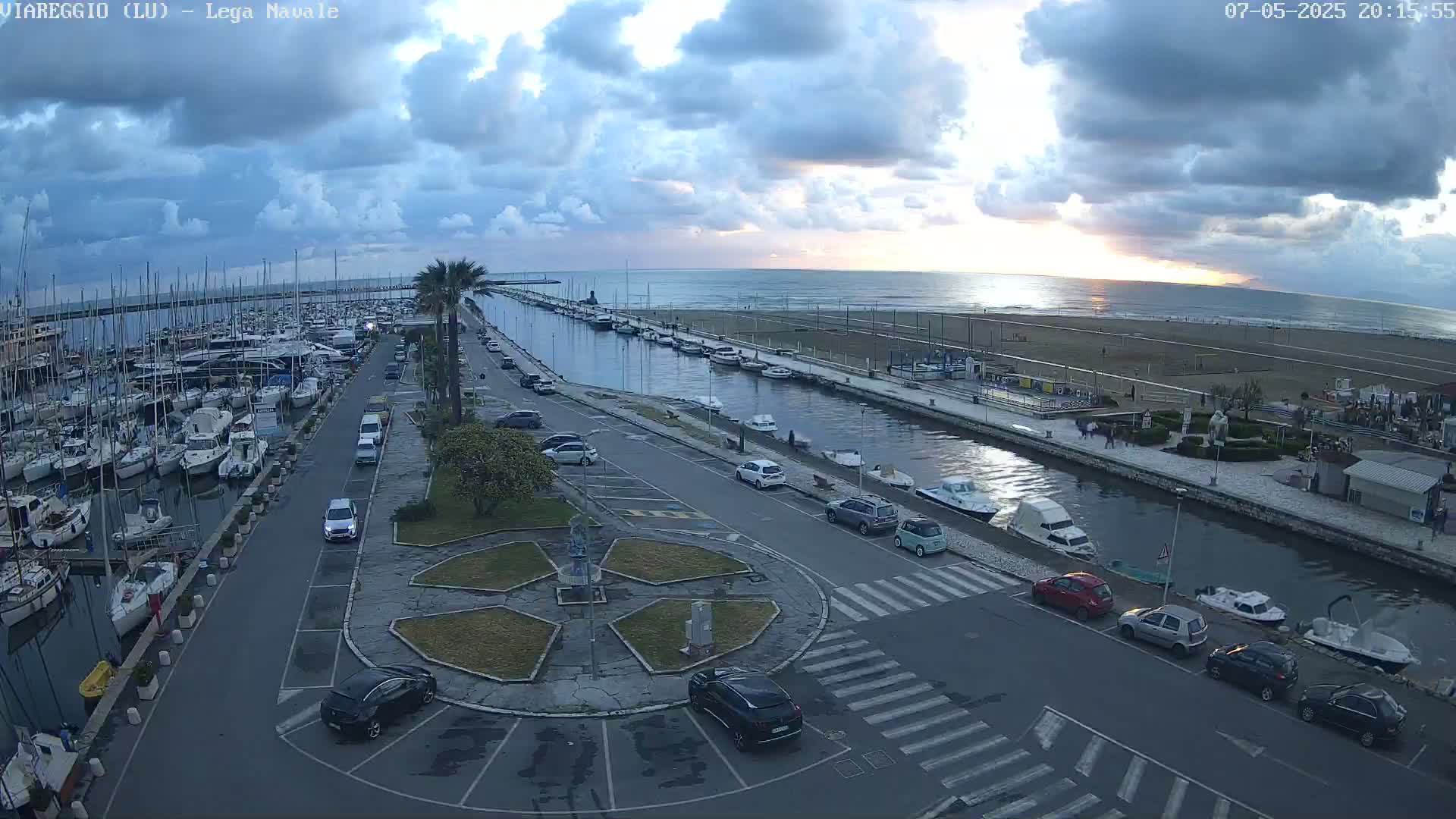 A marina filled with sailboats and motorboats is next to a canal and beach under a partly cloudy sunset sky.