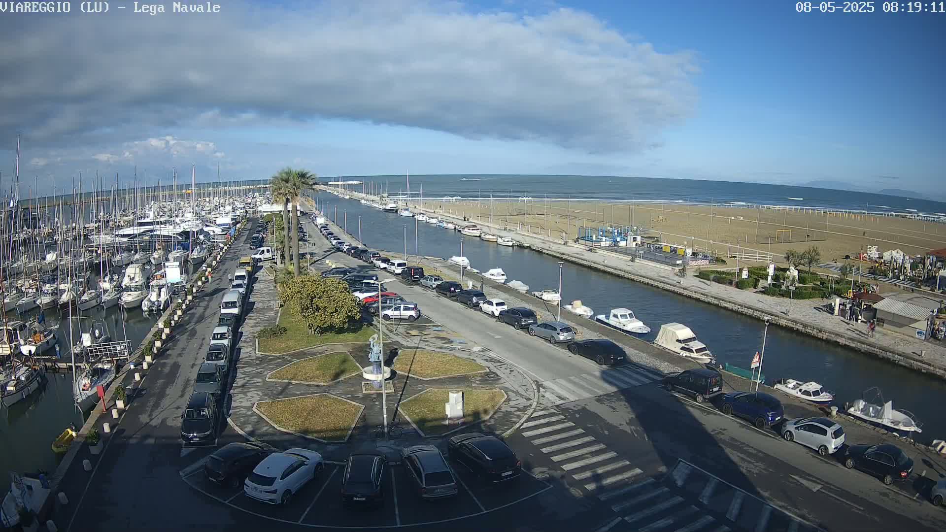 A marina filled with boats is adjacent to a canal and a sandy beach under a partly cloudy sky.