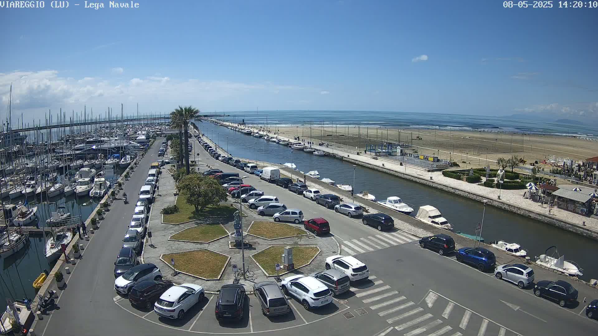 A marina filled with boats is next to a canal that leads to a sandy beach under a sunny sky.