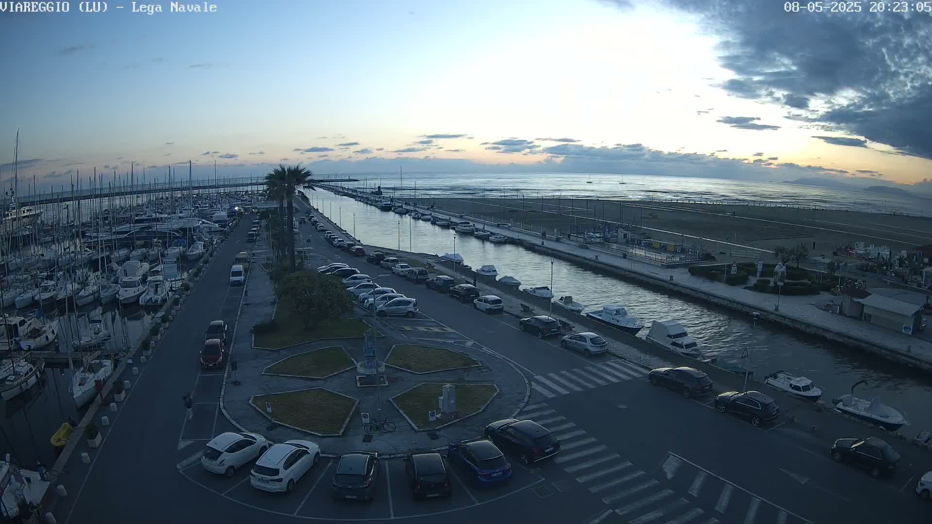 A marina filled with sailboats and motorboats is situated next to a canal that leads to a beach under a partly cloudy twilight sky.