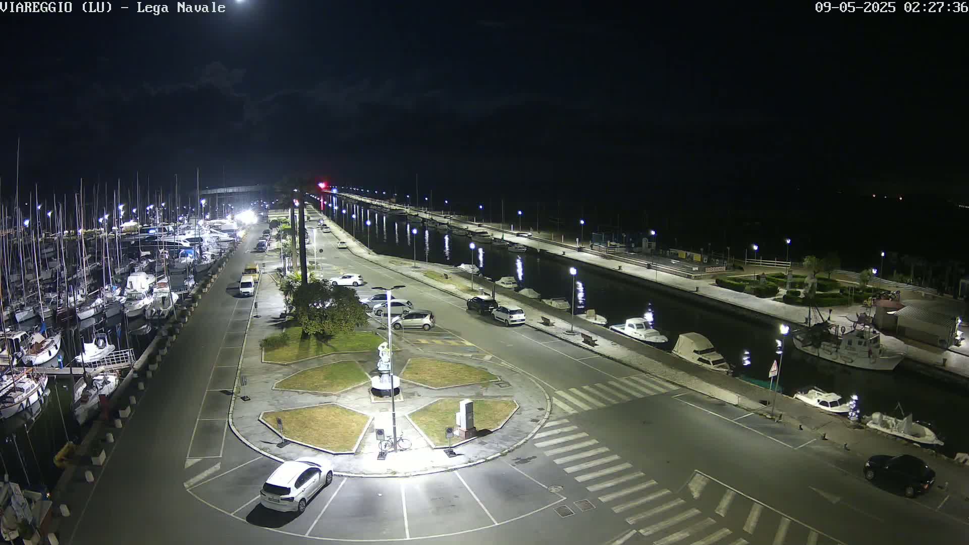 A nighttime aerial view of a marina with docked boats, cars parked along a roadway, and a small plaza in the center, under a mostly clear sky.
