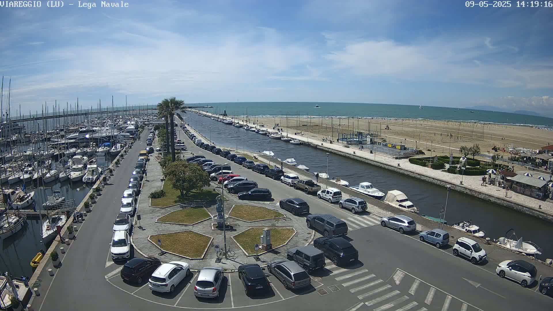 A marina filled with boats is adjacent to a canal and a sandy beach on a sunny day.