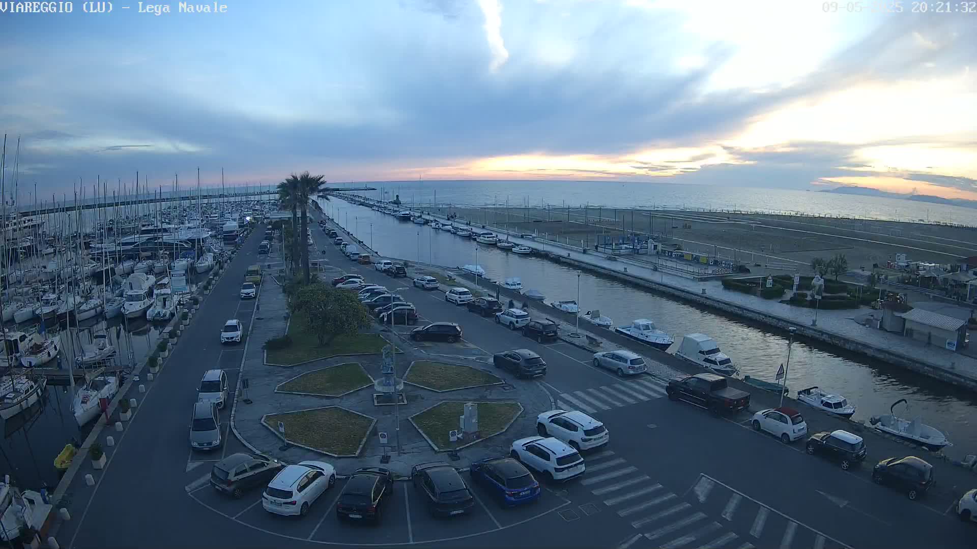 A marina filled with boats sits next to a canal lined with parked cars, all under a partly cloudy sunset sky.