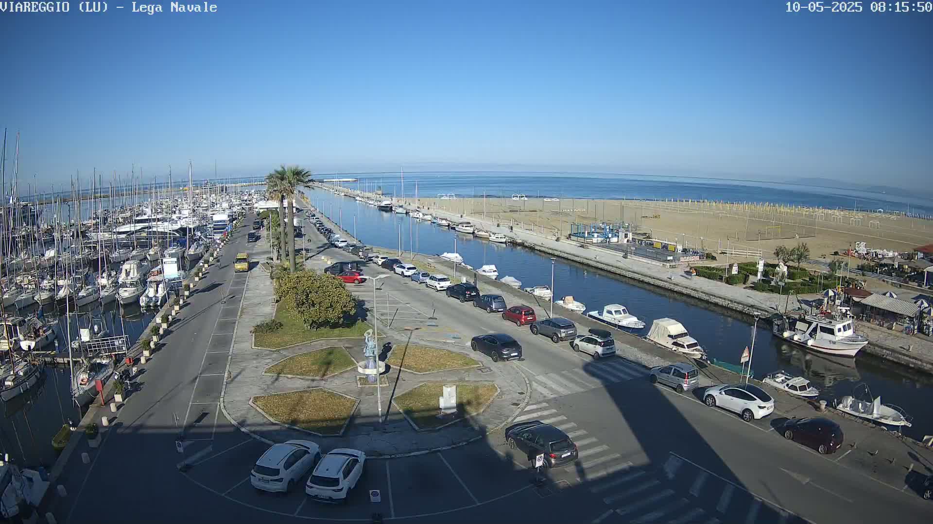 A marina filled with boats and surrounded by cars parked in a lot next to a canal leading to a sandy beach under a clear blue sky.