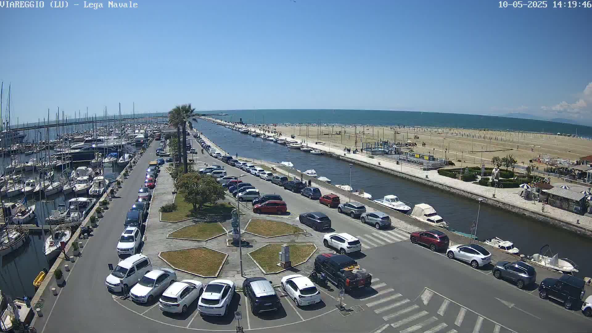 A marina filled with boats is adjacent to a canal lined with parked cars, which leads to a sandy beach under a clear blue sky.