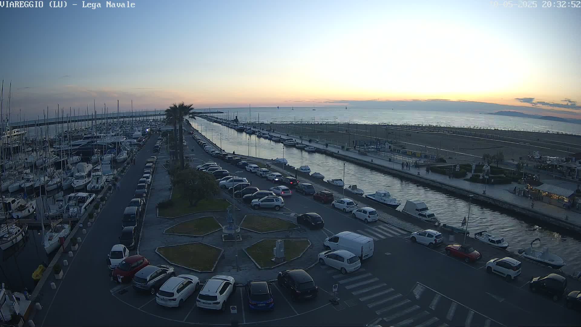 A marina filled with boats is adjacent to a calm canal and a beach at sunset, with cars parked along the waterfront.