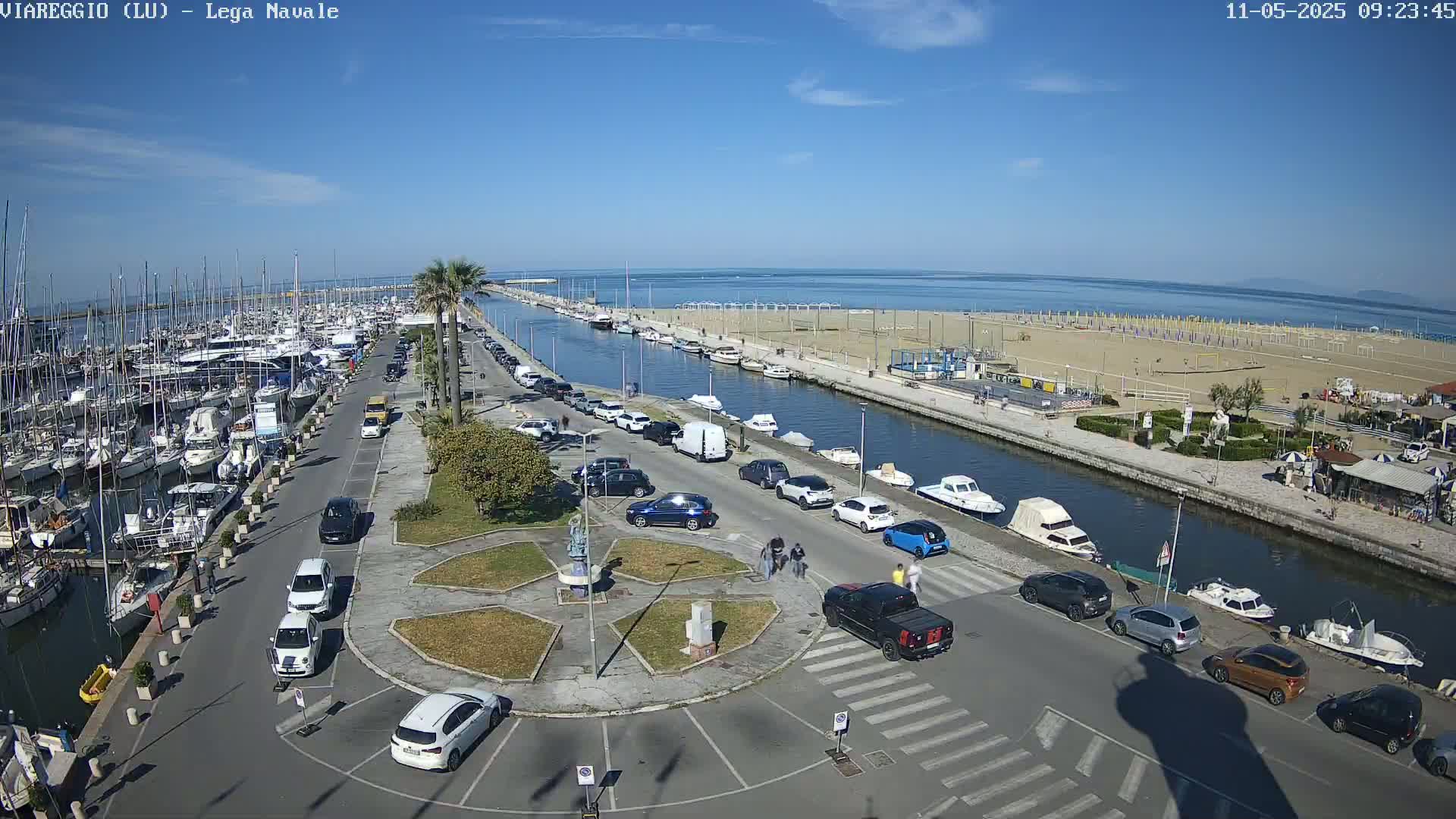 A marina filled with boats is adjacent to a sandy beach and calm ocean under a sunny sky.
