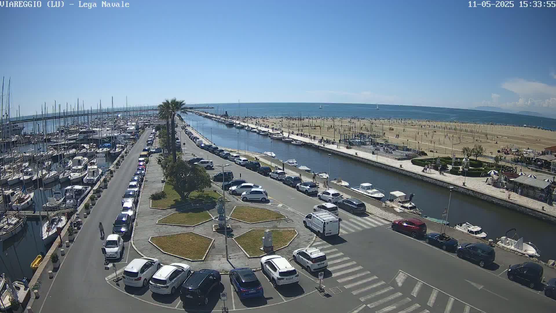 A marina filled with boats, a canal, a sandy beach with people, and a parking lot with cars are seen on a sunny day.