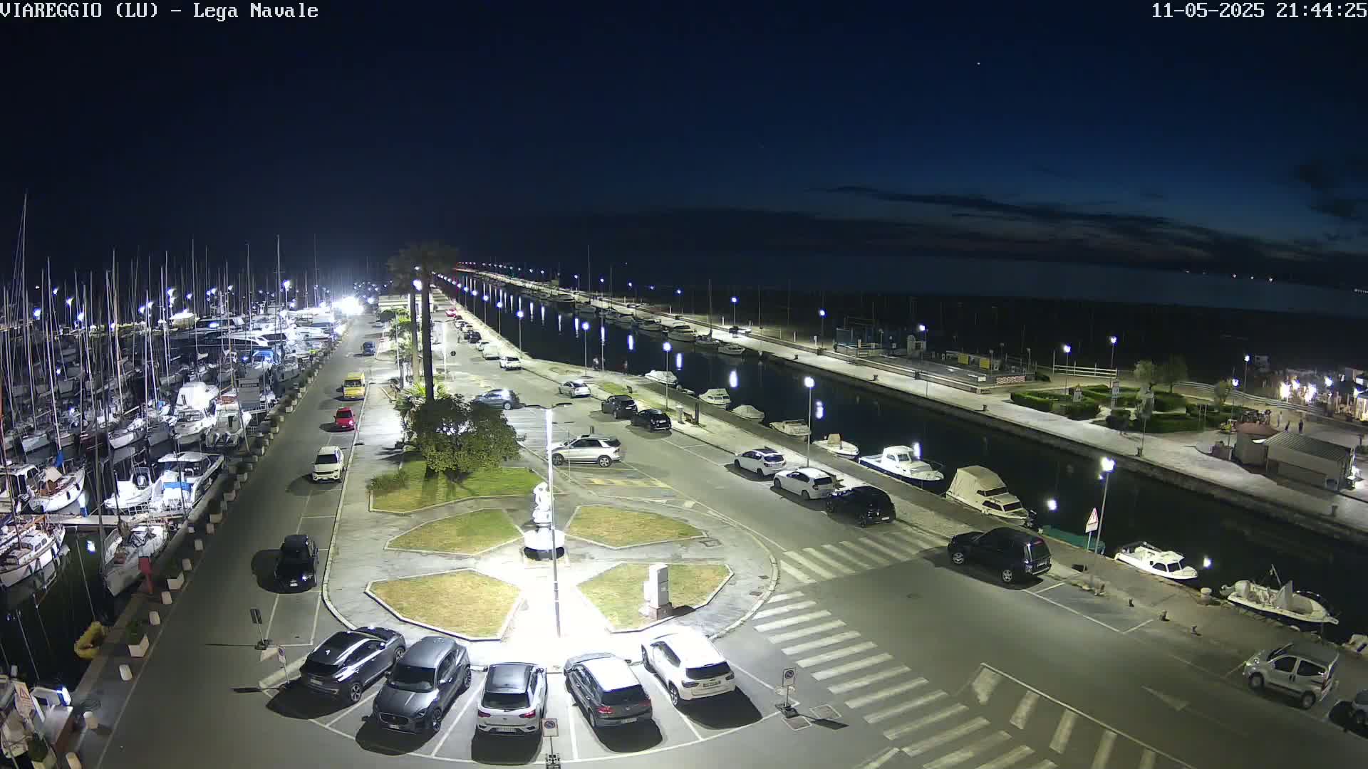 A nighttime aerial view of a marina with boats docked, cars parked in a lot, and a calm canal separating the parking area from the waterfront.