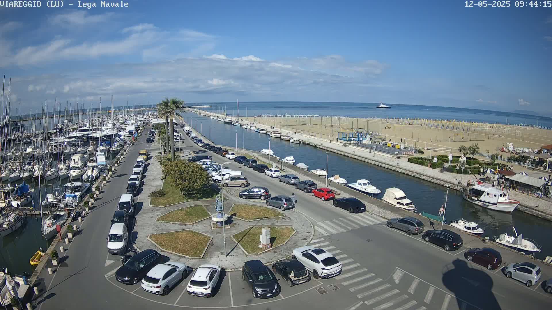 A marina filled with boats is adjacent to a sandy beach and calm ocean under a mostly sunny sky, with cars parked along the roadway that separates them.