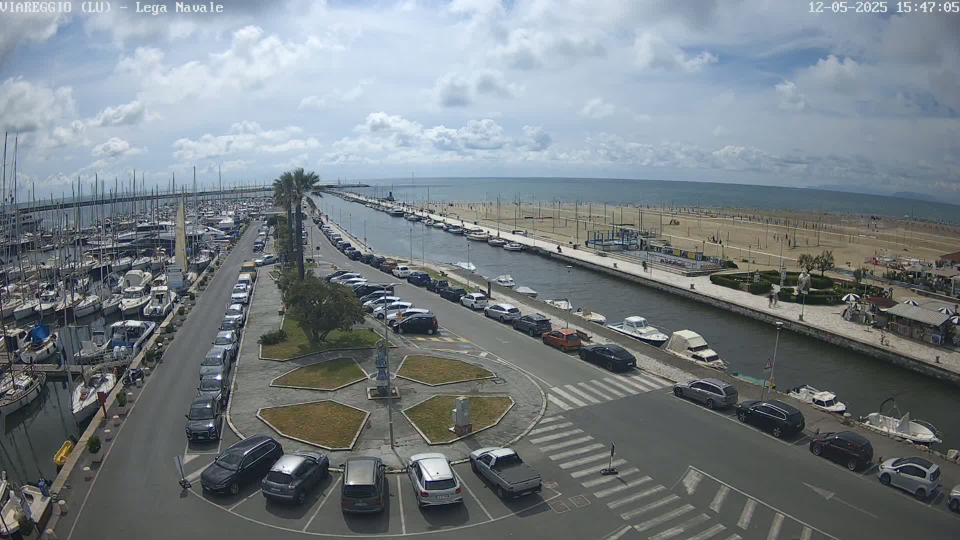 A marina filled with boats, adjacent to a sandy beach and a canal lined with parked cars under a partly cloudy sky.