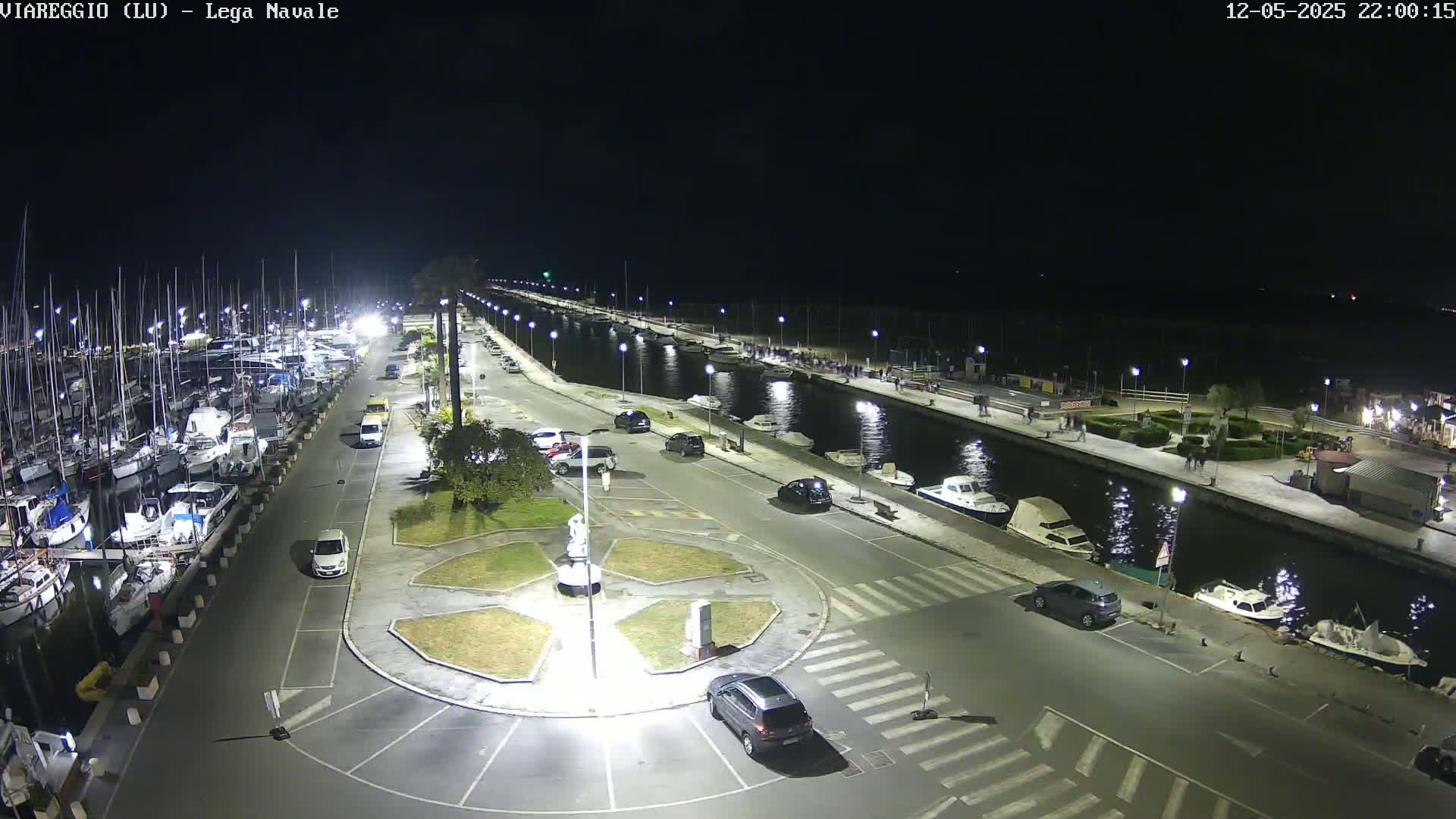A nighttime aerial view shows a marina filled with boats, a canal with several more boats, and a paved area with cars parked alongside, all illuminated by streetlights.
