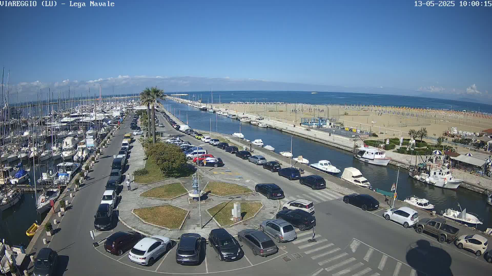 A marina filled with boats is adjacent to a sandy beach with numerous umbrellas, all under a clear blue sky.