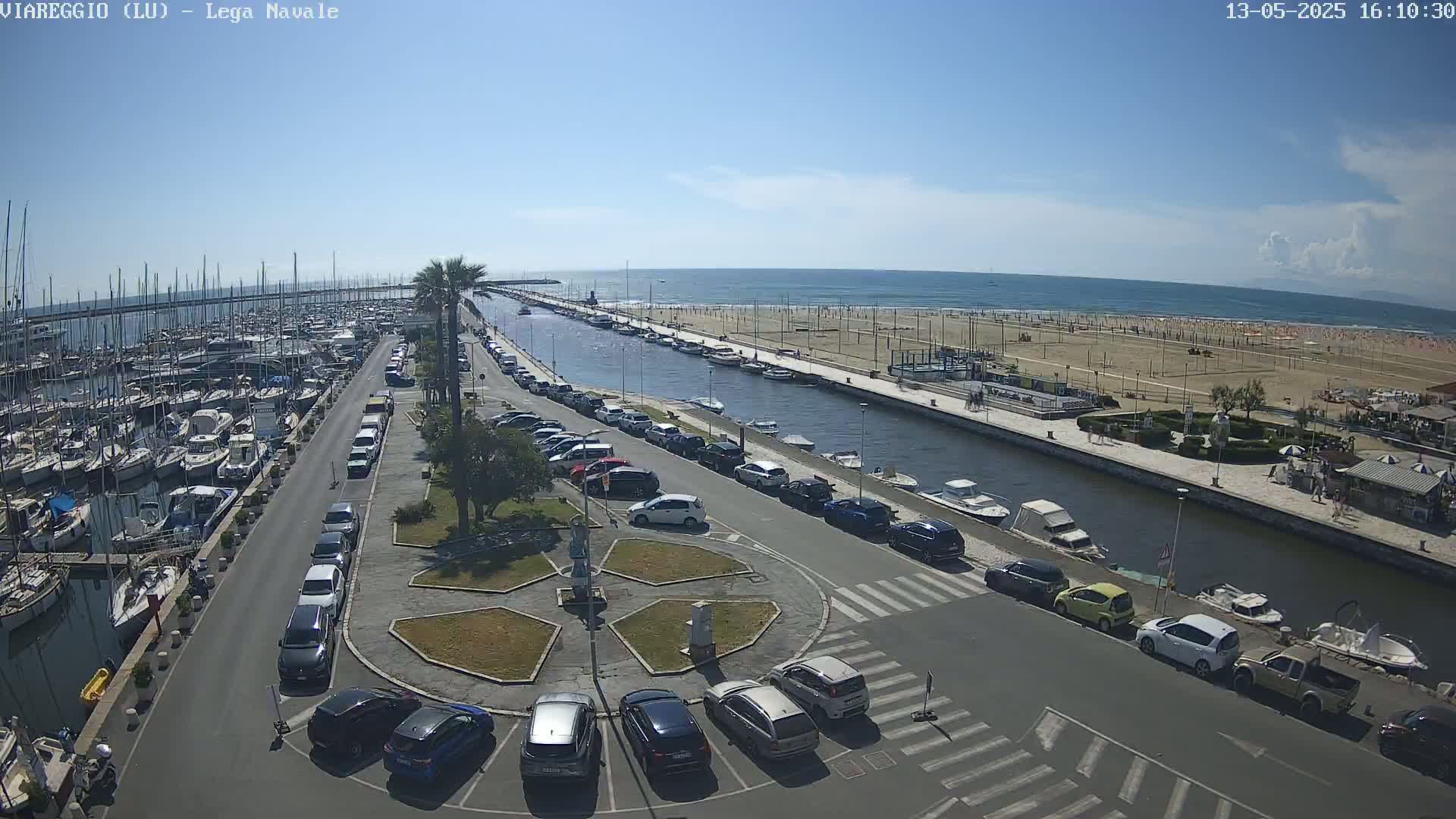 A marina filled with boats is adjacent to a canal, which runs alongside a sandy beach under a clear blue sky.