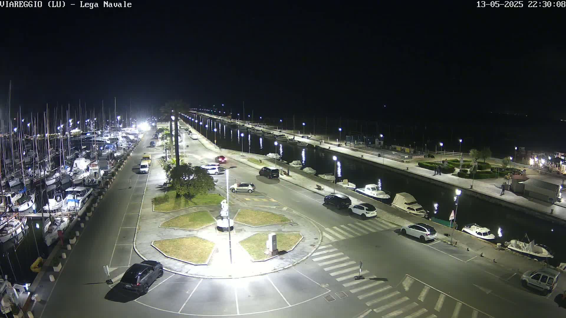 A nighttime aerial view of a marina with many boats docked, cars parked along a paved walkway, and a central roundabout.