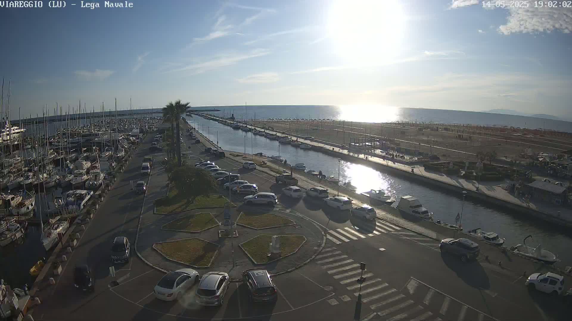 A sunny harbor scene shows many boats docked, cars parked along a canal, and a sandy beach in the distance.
