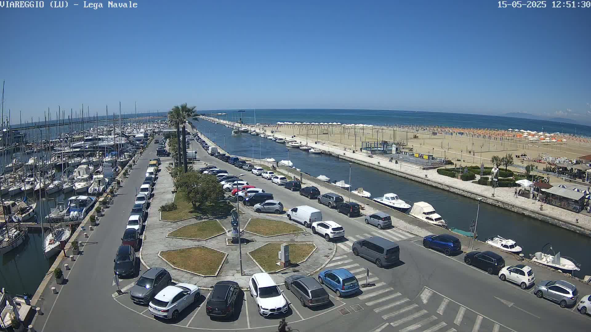 A marina filled with boats is adjacent to a canal, which leads to a sandy beach with numerous umbrellas and people under a clear blue sky.