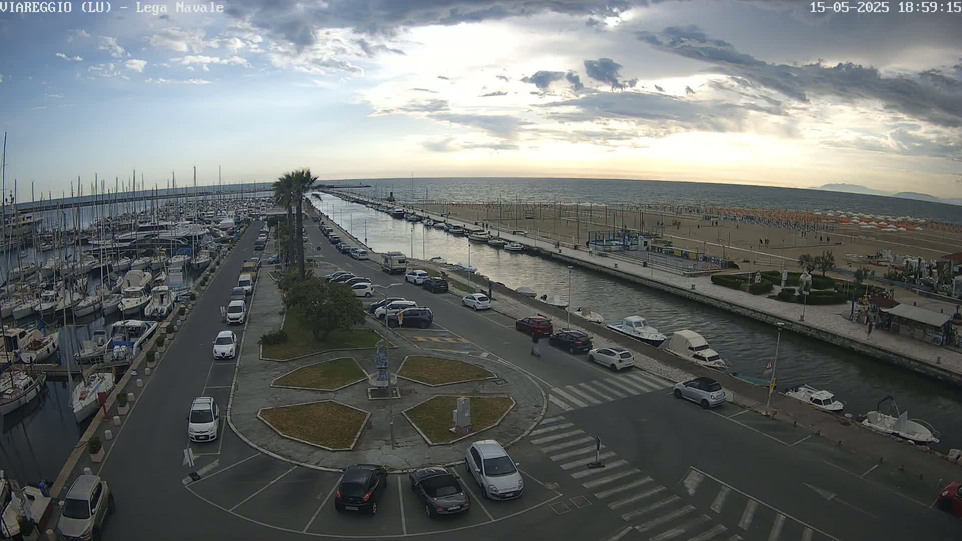 A harbor filled with boats is adjacent to a beach with numerous umbrellas and people, all under a partly cloudy sky.