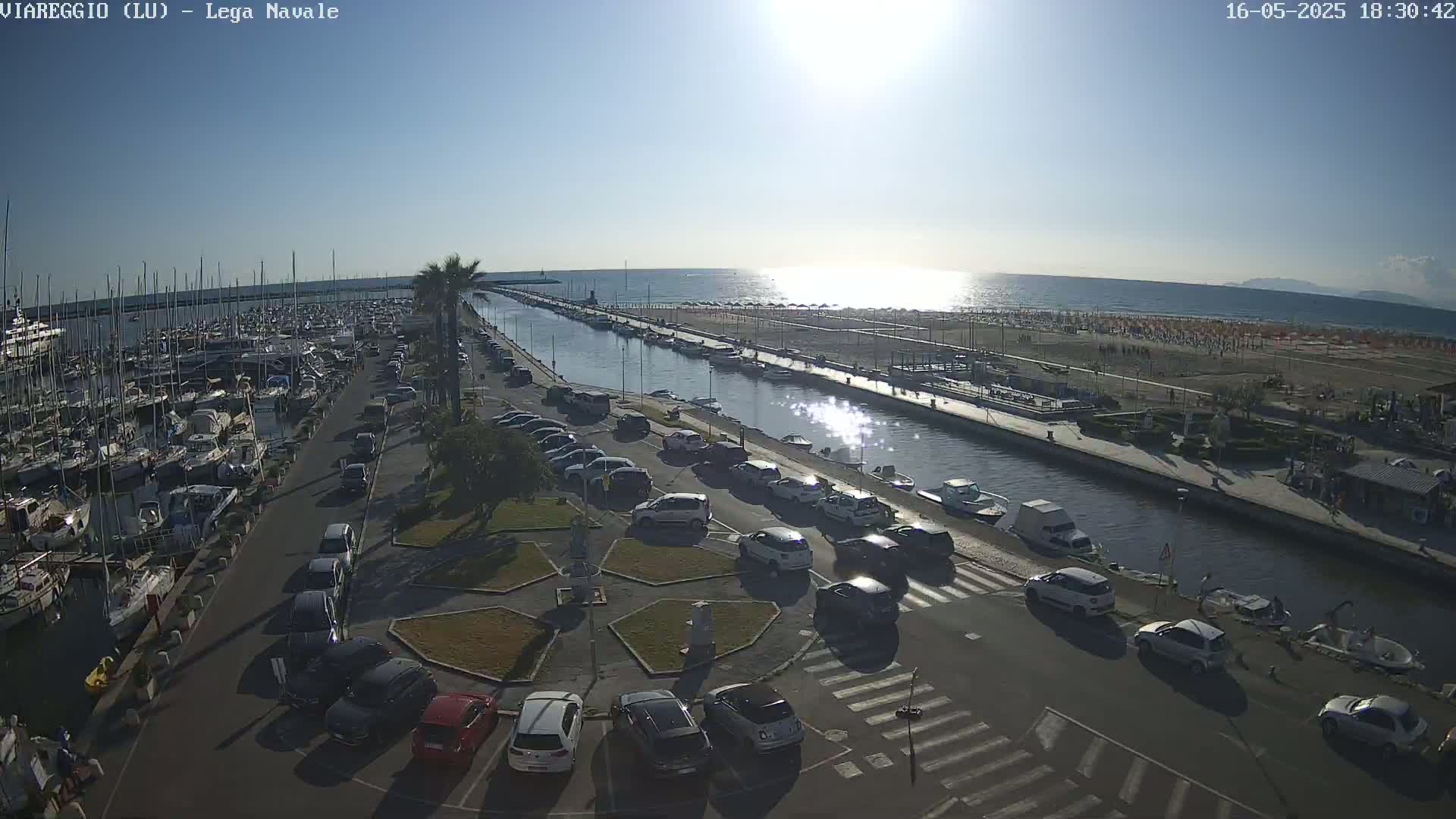 A harbor filled with boats is next to a canal with parked cars alongside it, all bordering a sunny beach with people on it under a clear sky.