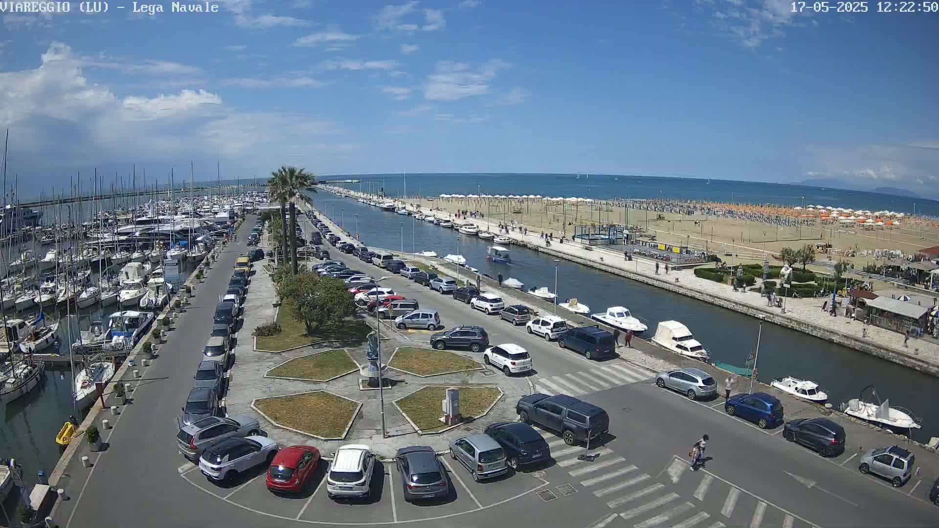 A marina filled with boats, a canal, a beach with numerous umbrellas, a parking lot full of cars, and a sunny day.
