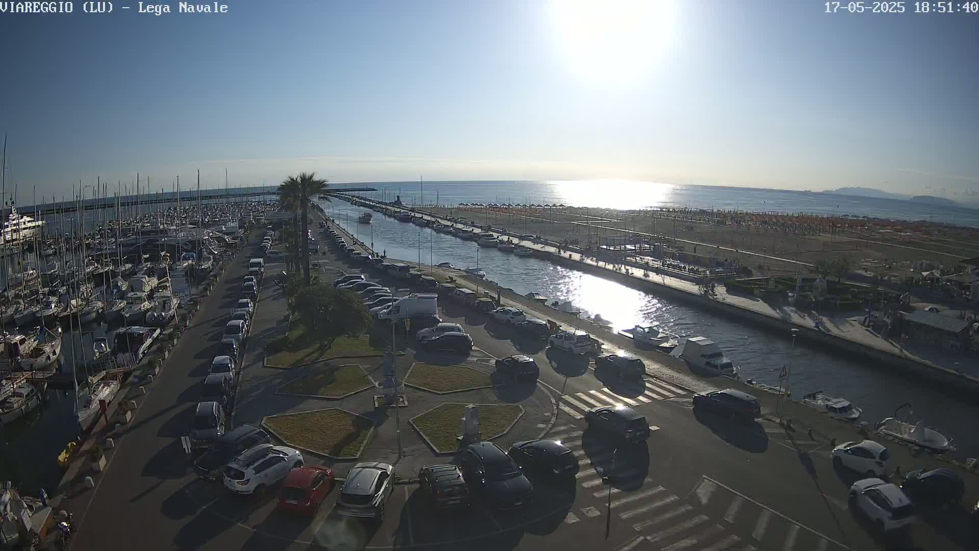 A sunny day reveals a harbor filled with boats, a canal lined with parked cars and people on a beach, and a clear blue ocean in the background.