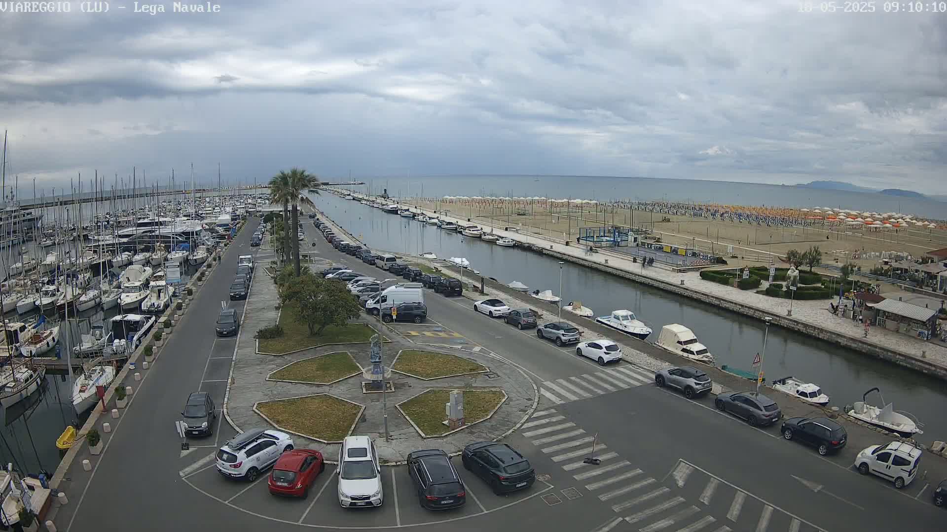 A marina filled with boats is next to a canal that leads to a beach covered in umbrellas under an overcast sky.