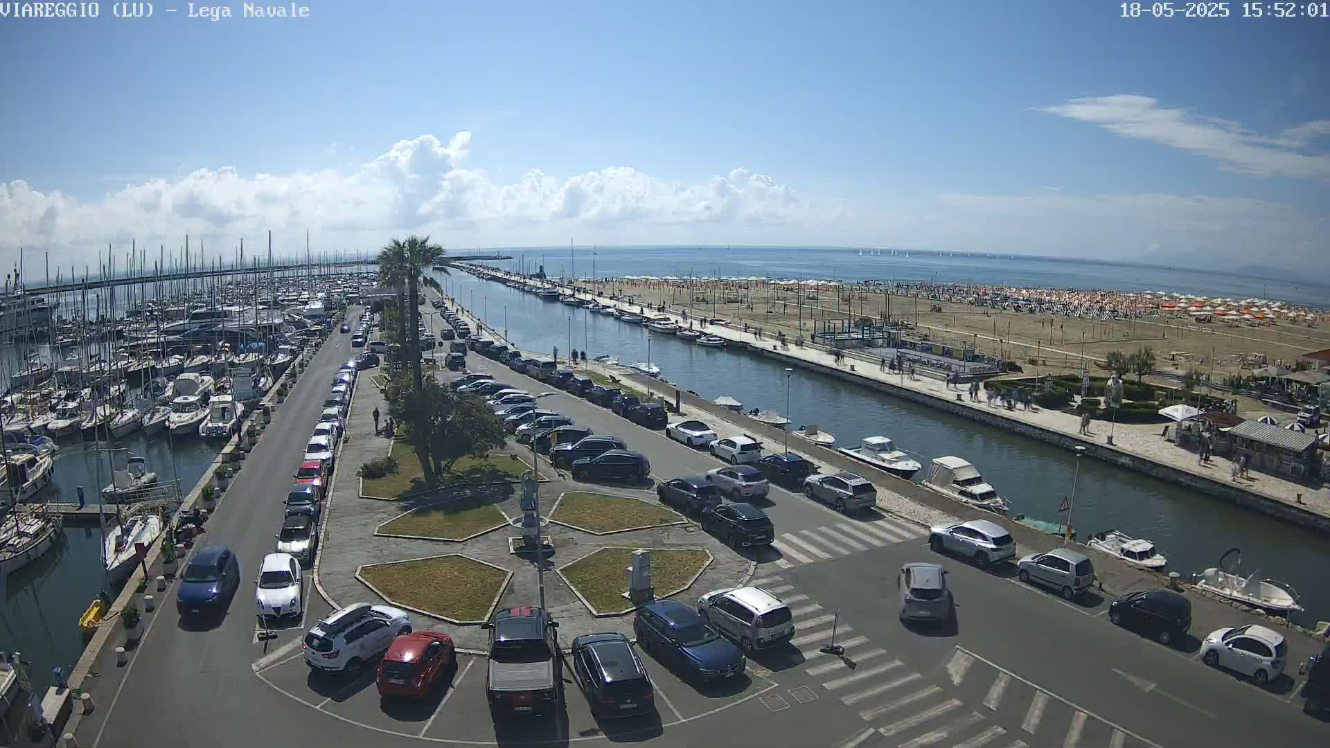 A marina filled with boats is adjacent to a canal that leads to a beach crowded with people under a partly cloudy sky.