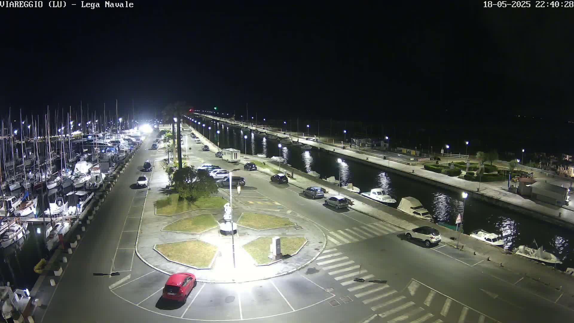 A nighttime aerial view of a marina with docked boats, cars parked along a paved walkway, and a canal reflecting lights.