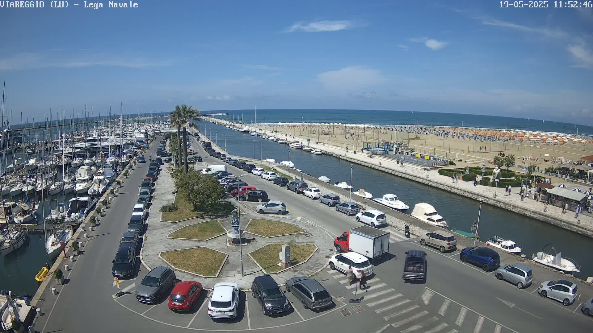 A marina filled with boats is adjacent to a parking lot and a canal leading to a sandy beach with numerous umbrellas and people under a sunny sky.