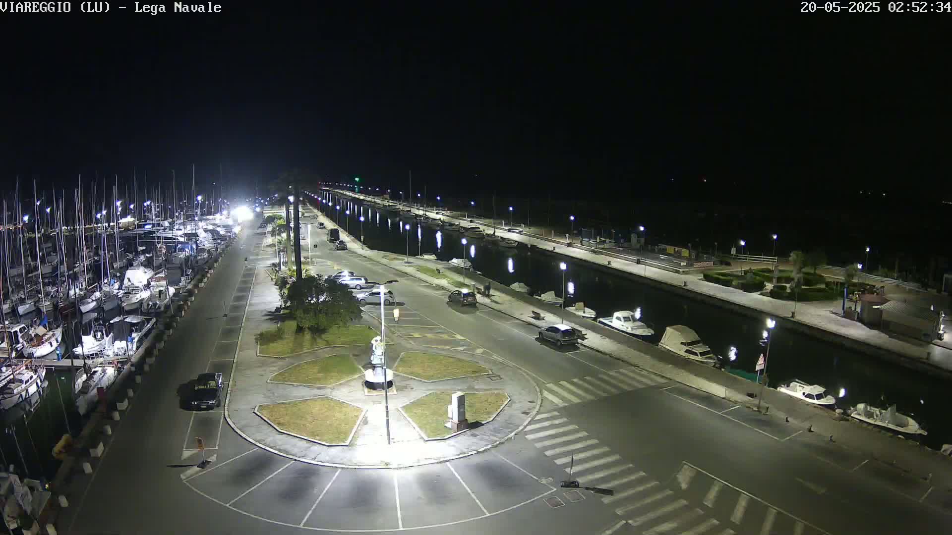 A nighttime aerial view of a marina with numerous boats docked alongside a paved waterfront area containing parked cars and streetlights.