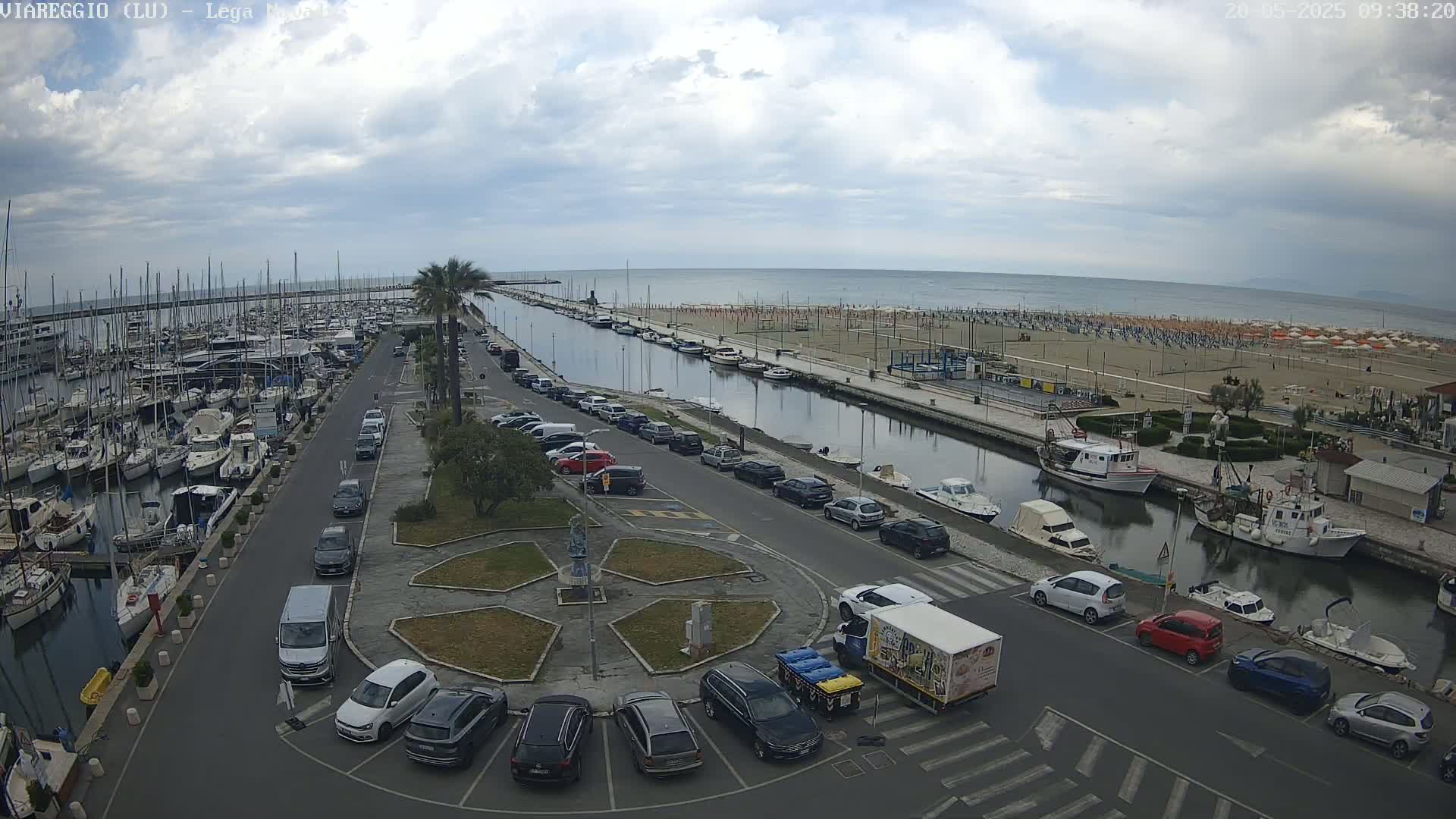 A marina filled with boats, a canal, and a parking lot next to a sandy beach under a cloudy sky.