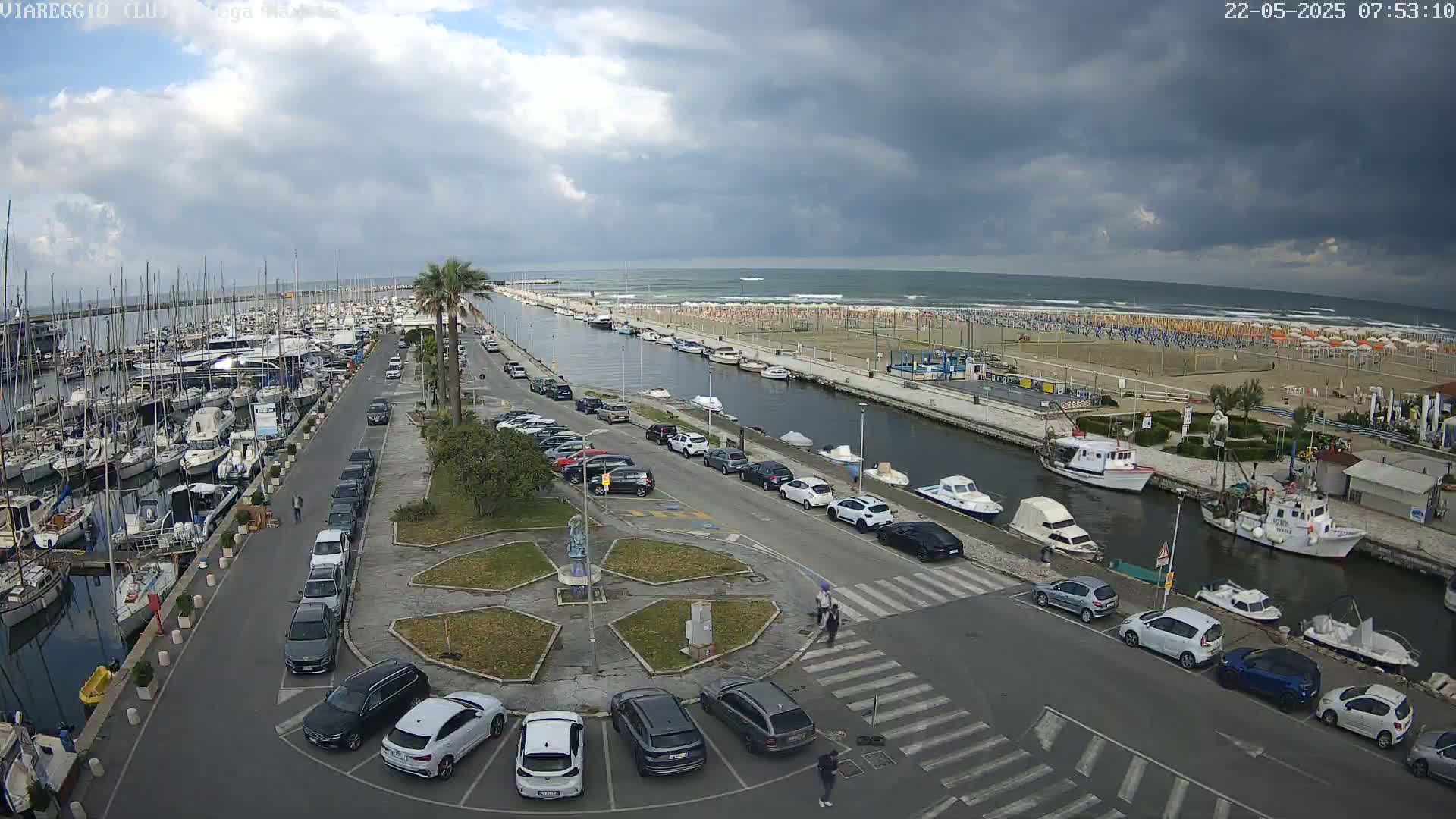 A marina filled with boats is next to a parking lot and a canal leading to a beach with numerous umbrellas under an overcast sky.
