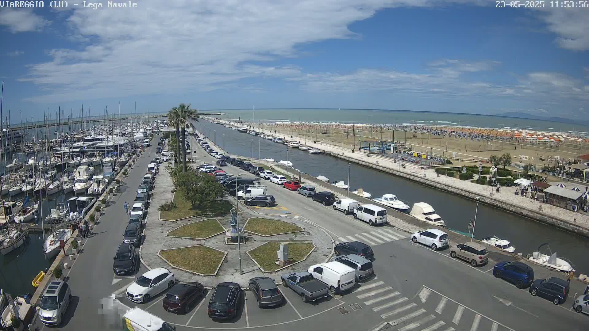 A marina filled with boats, next to a canal with parked cars, and a beach with people under sunny skies.