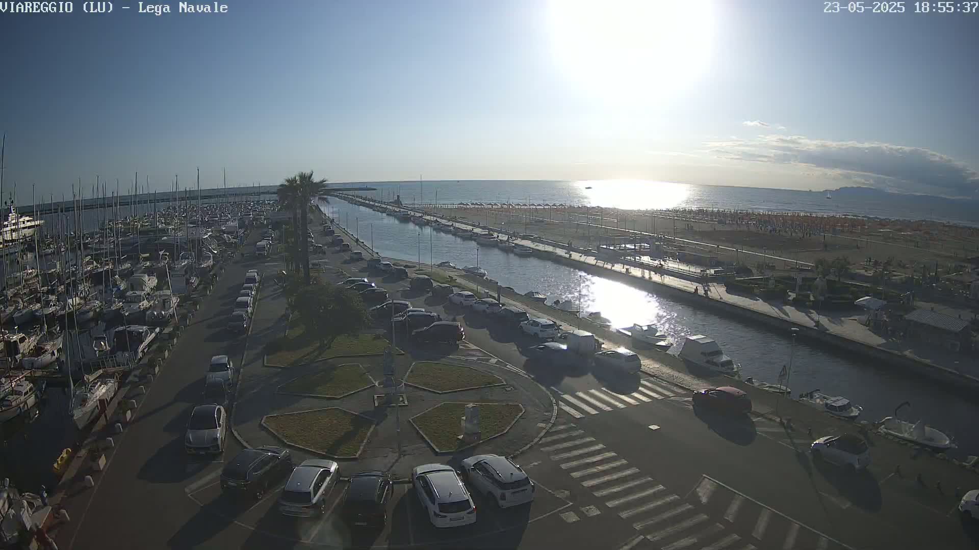 A sunny day overlooking a marina filled with boats, a canal, a beach with umbrellas, and a parking lot with cars.