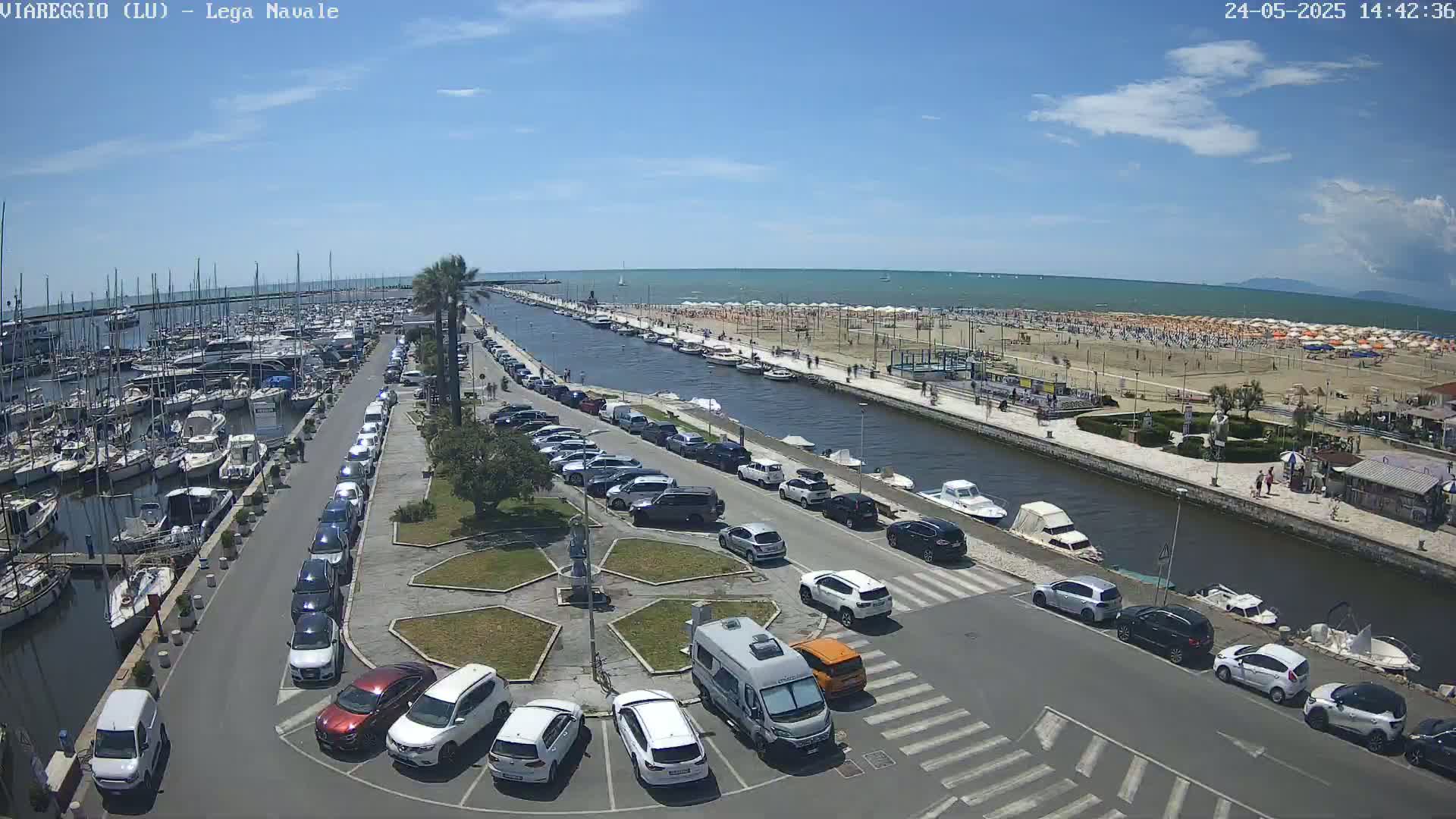 A marina filled with boats is next to a canal that leads to a sandy beach crowded with people under a sunny sky.