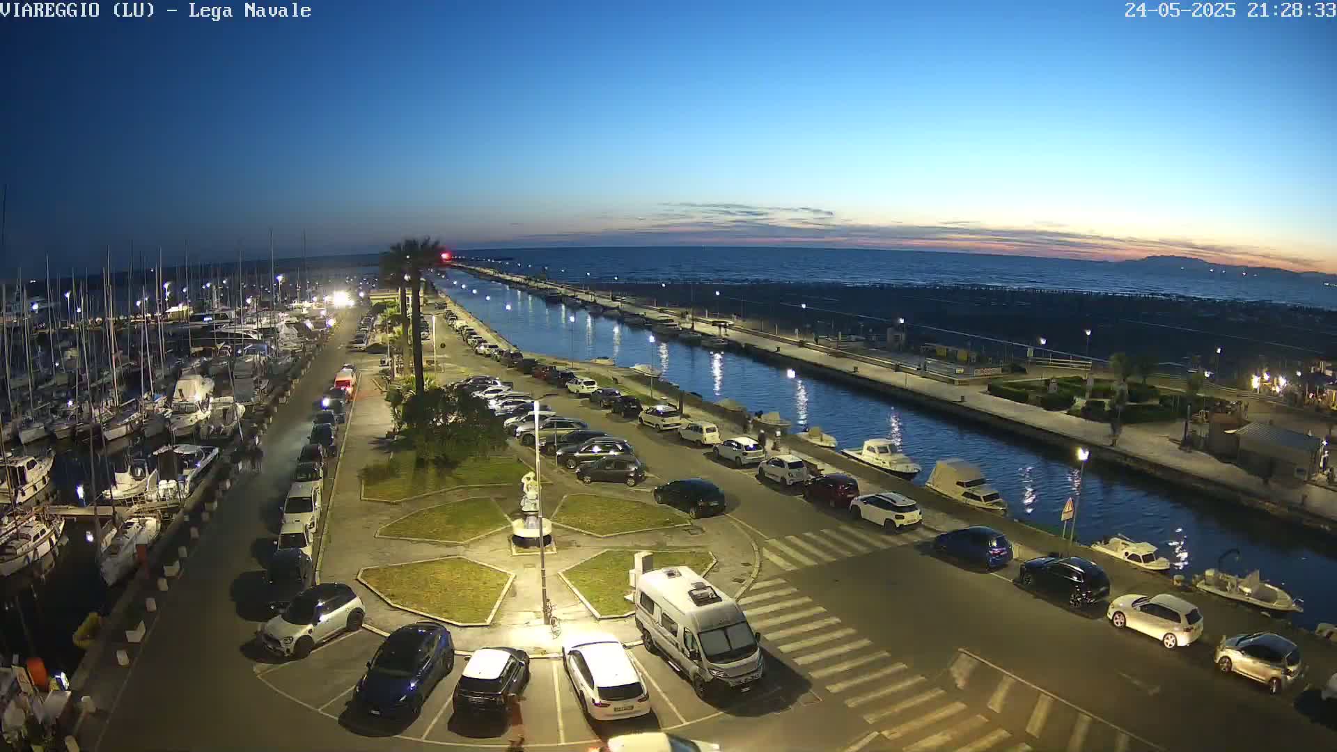 A marina at twilight, with cars parked along a waterfront promenade next to a canal, and boats moored in the harbor under a clear sky.