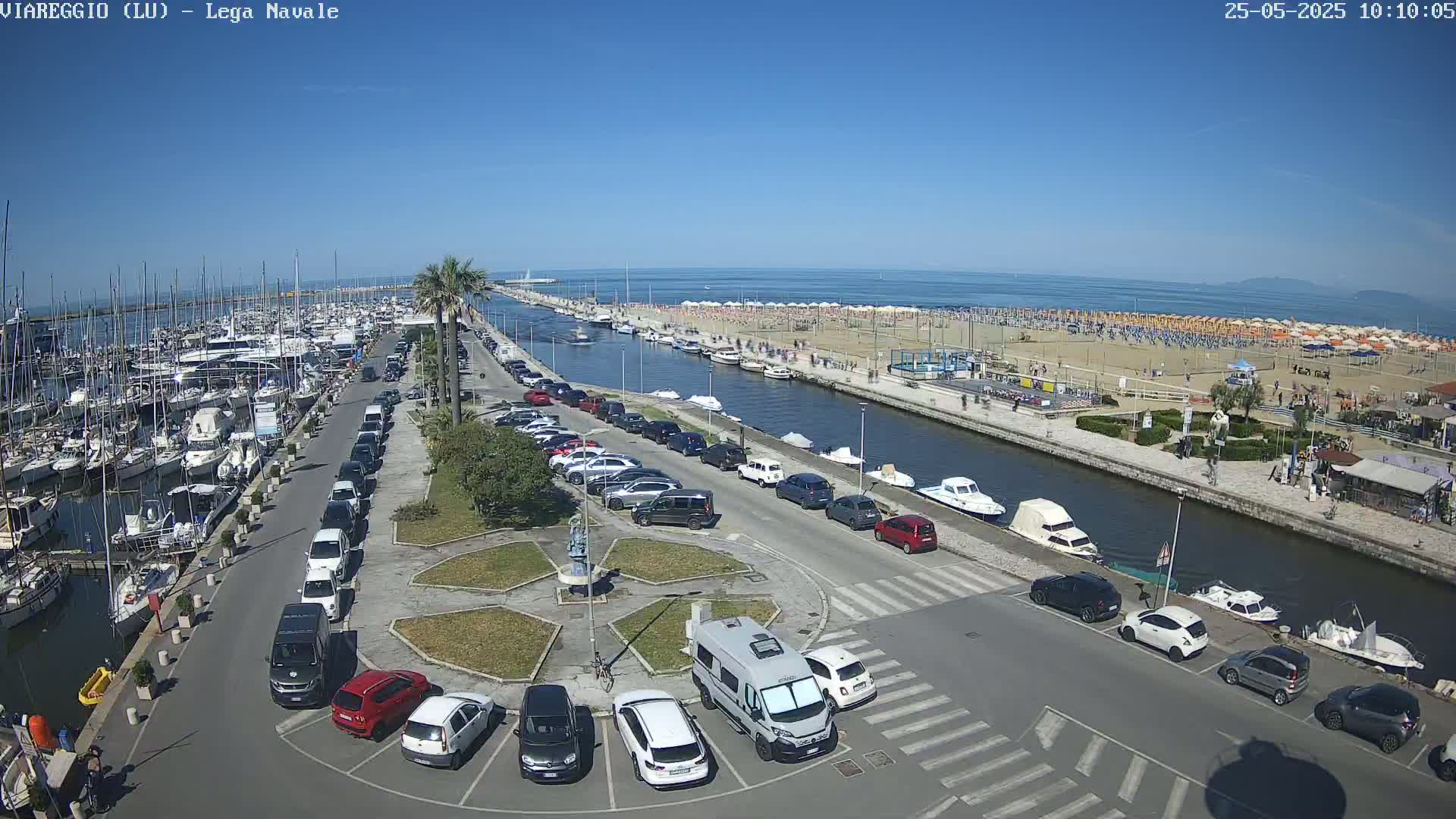 An aerial view of a harbor filled with boats, adjacent to a sandy beach with numerous umbrellas and people, under sunny skies, with a parking lot separating the two.