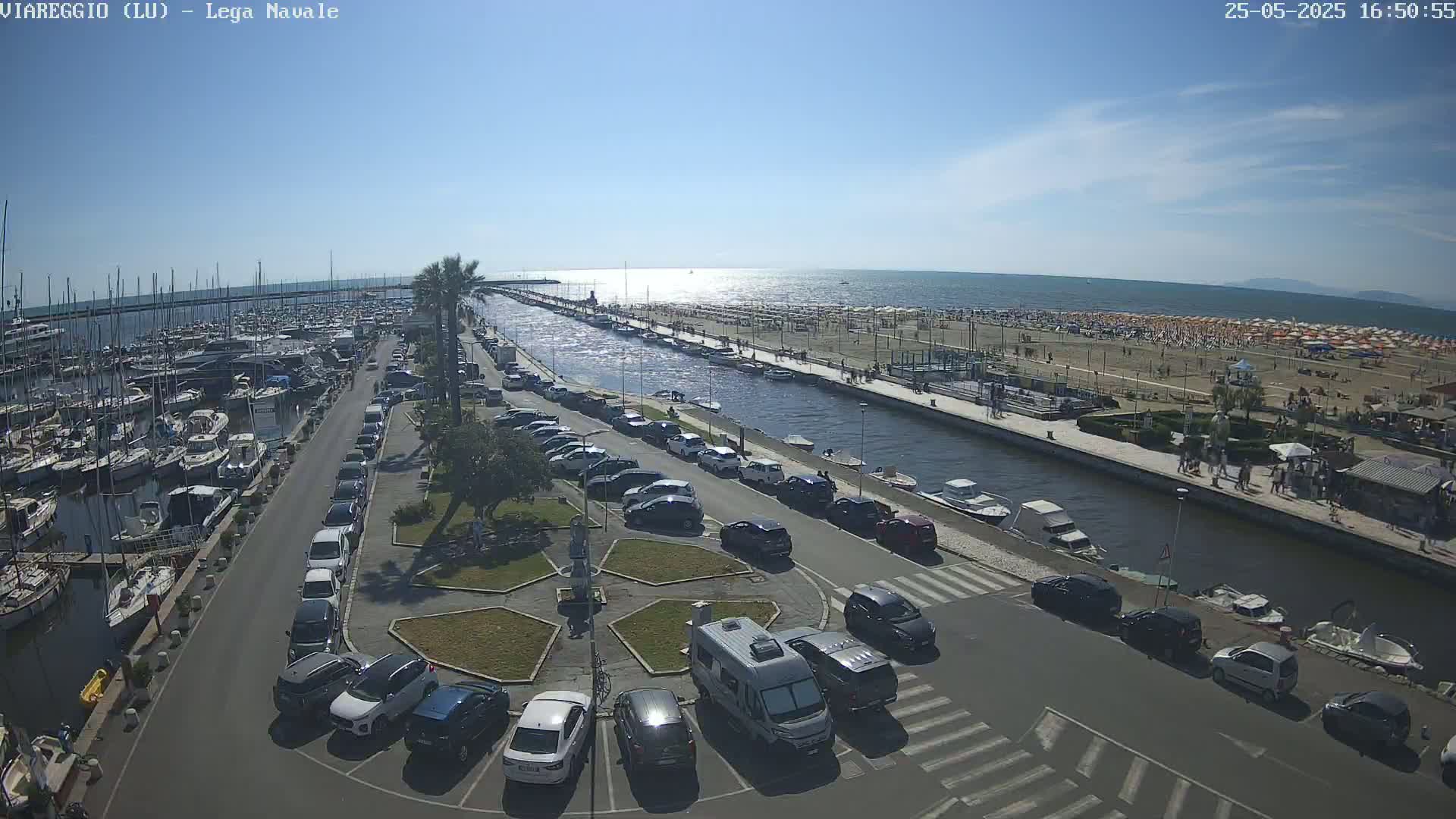A sunny day shows a marina filled with boats, a canal lined with parked cars leading to a crowded beach with numerous umbrellas.