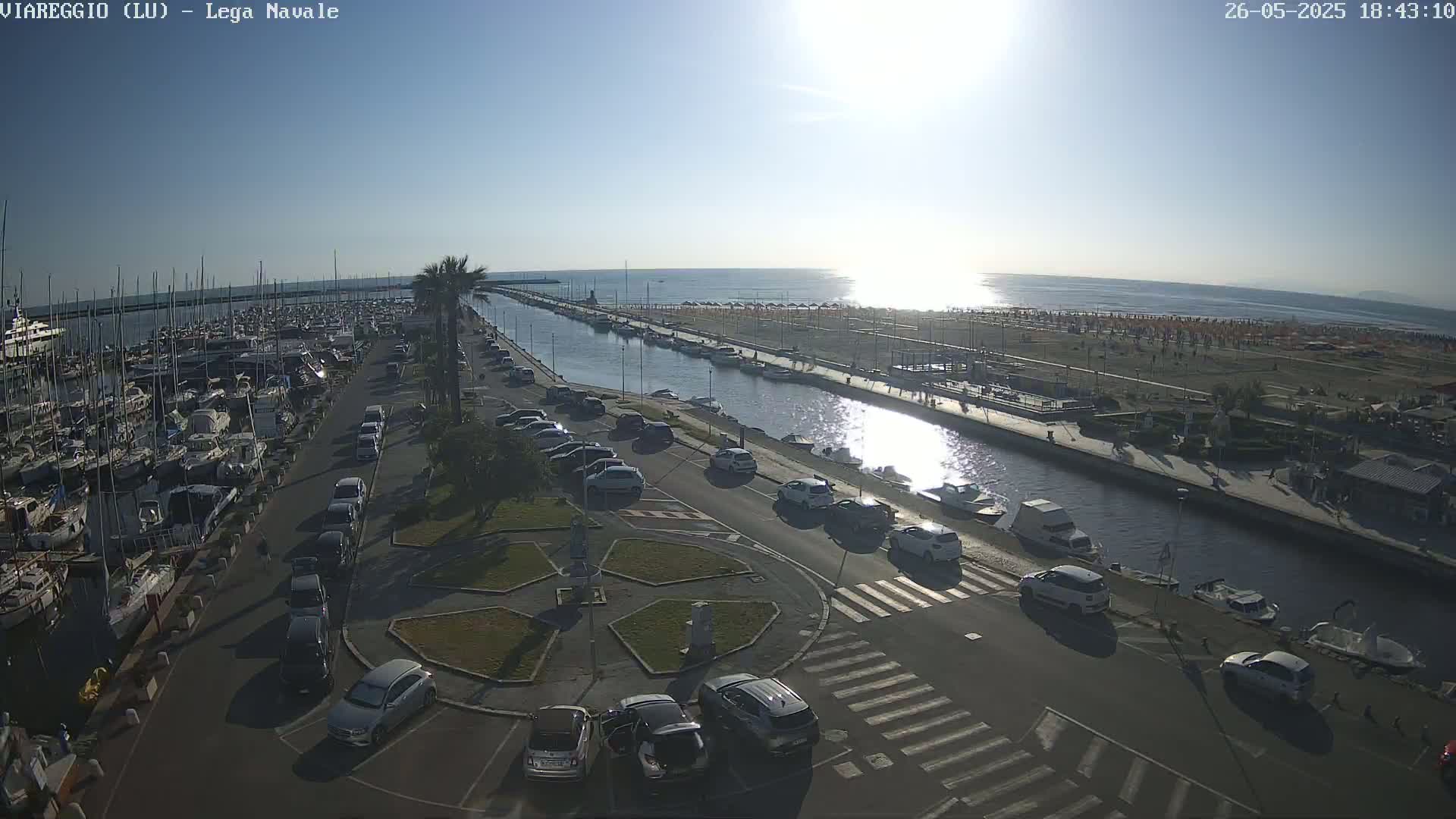 A marina filled with boats is next to a canal that leads to a beach on a sunny day.
