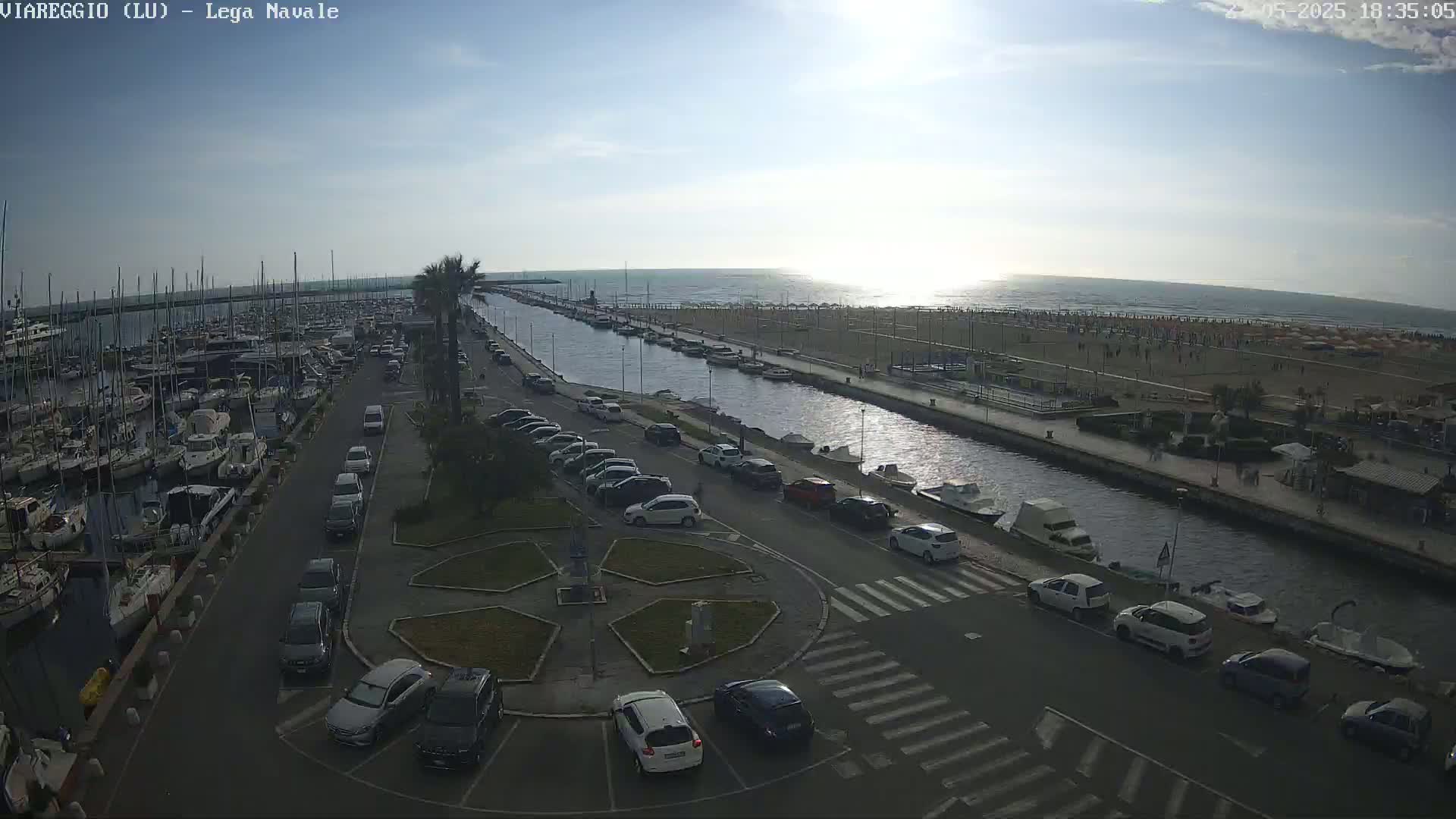 A marina filled with boats is next to a canal that leads to a beach with people on it under a partly sunny sky.