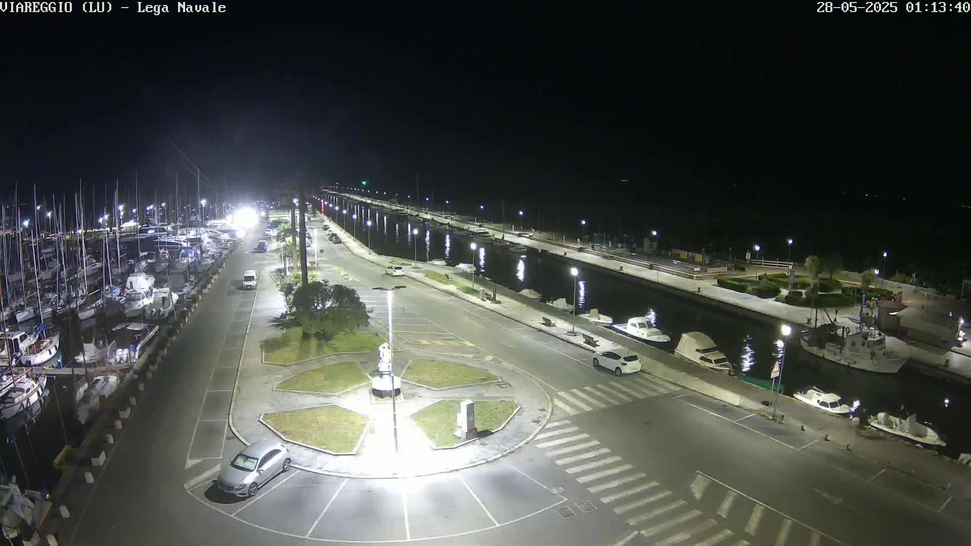 A nighttime, illuminated view of a marina with numerous boats docked along a canal, a roadway with parked cars, and a central roundabout.