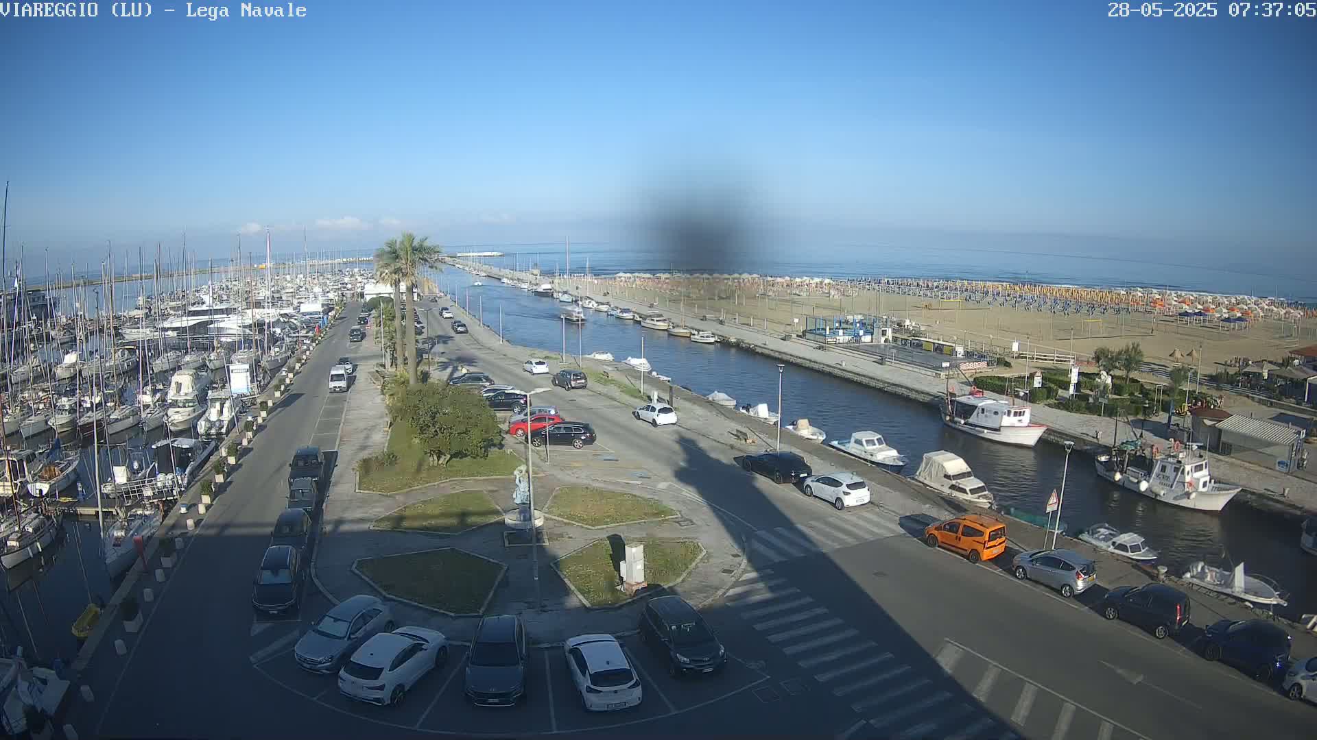 A marina filled with boats is adjacent to a canal and a beach with numerous umbrellas under a clear blue sky.