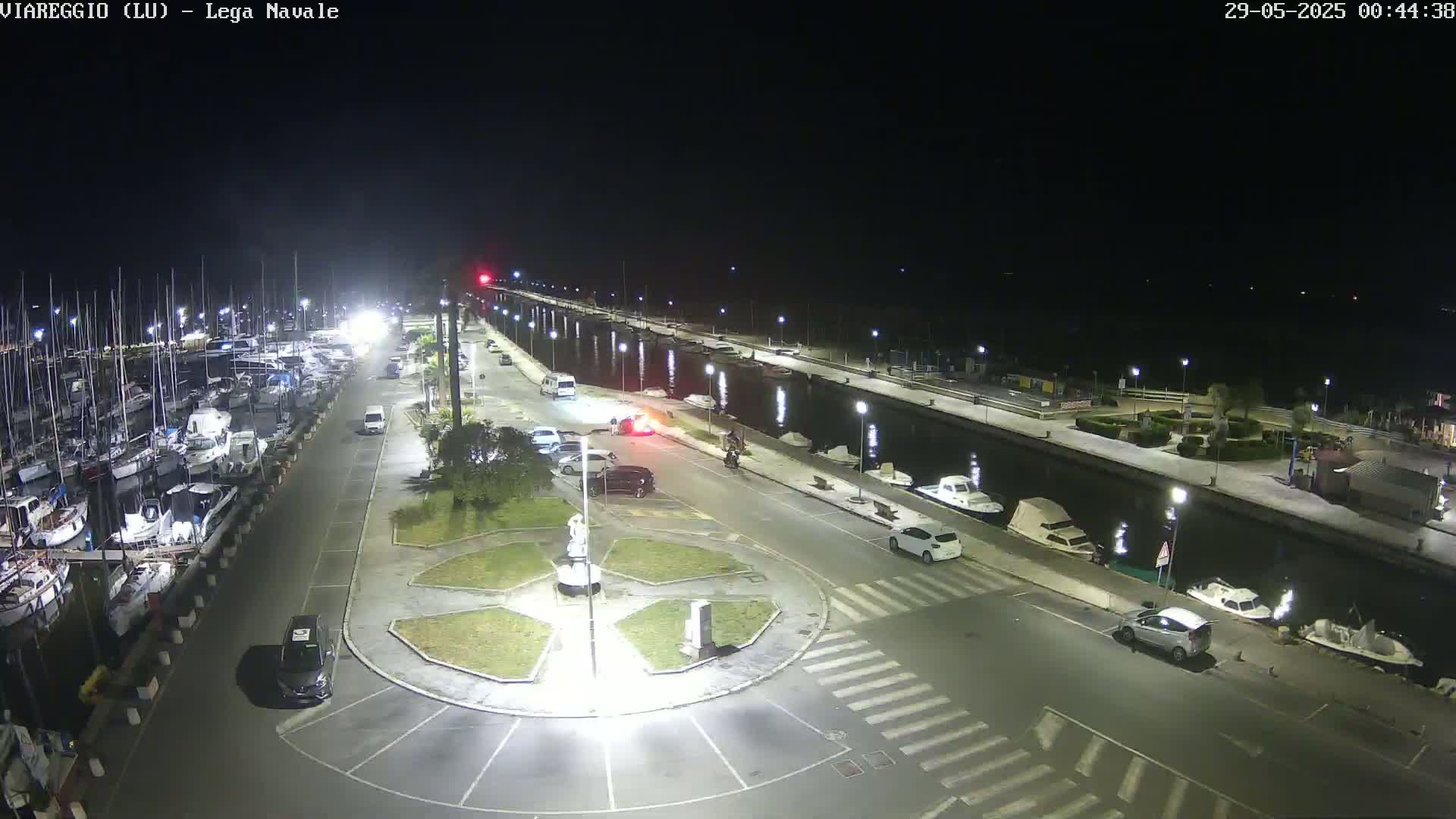 A nighttime aerial view shows a marina with numerous boats docked alongside a paved waterfront area containing several parked cars and a small, landscaped traffic circle.