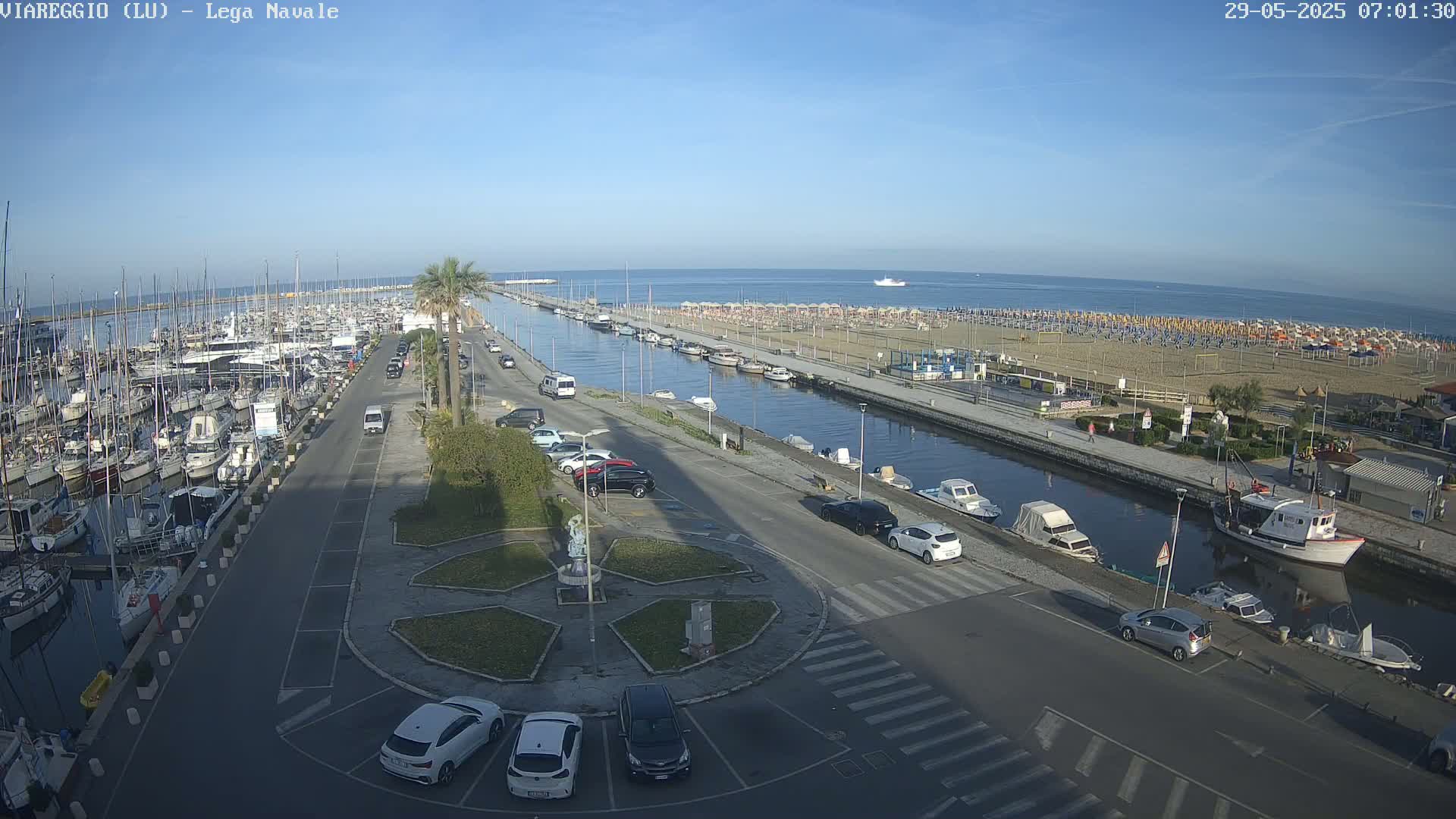 A marina filled with boats is next to a canal that leads to a sandy beach with numerous umbrellas and a calm ocean under a clear sky.