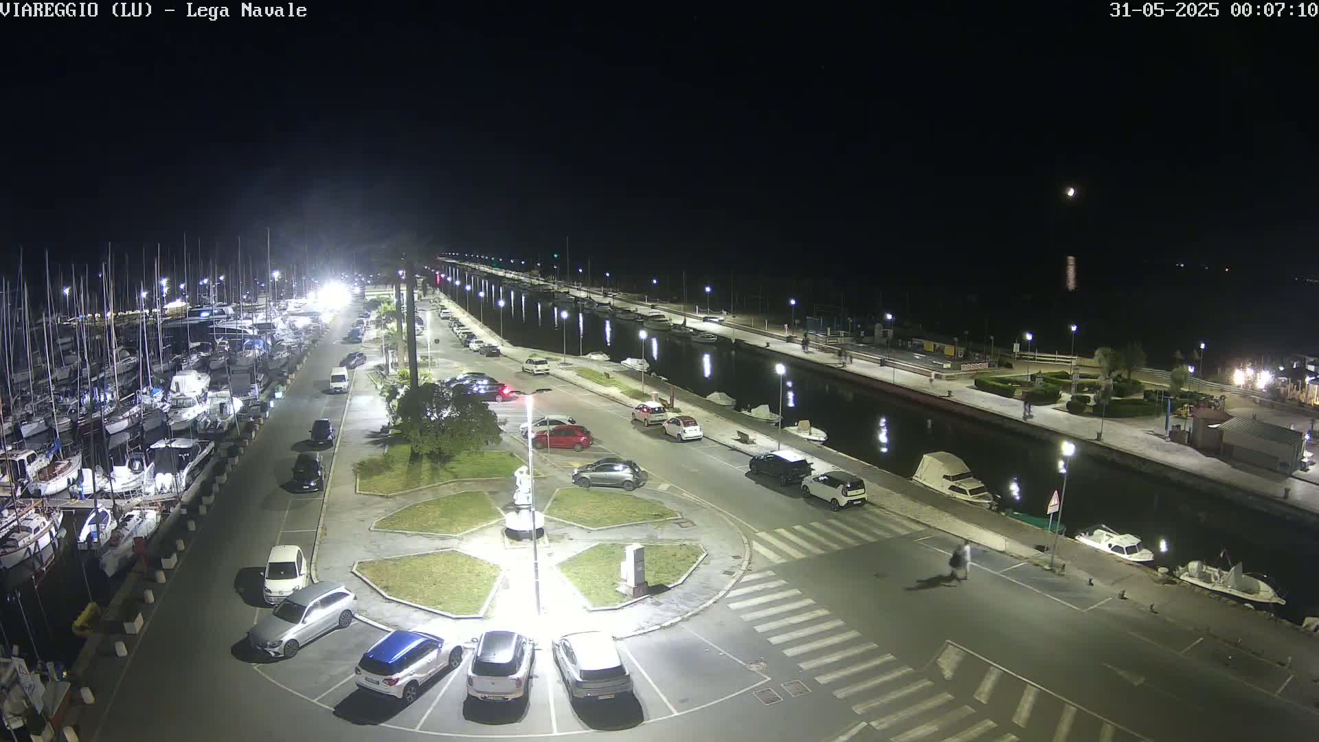 A nighttime aerial view of a marina with numerous boats docked, a paved area with parked cars, and a canal.