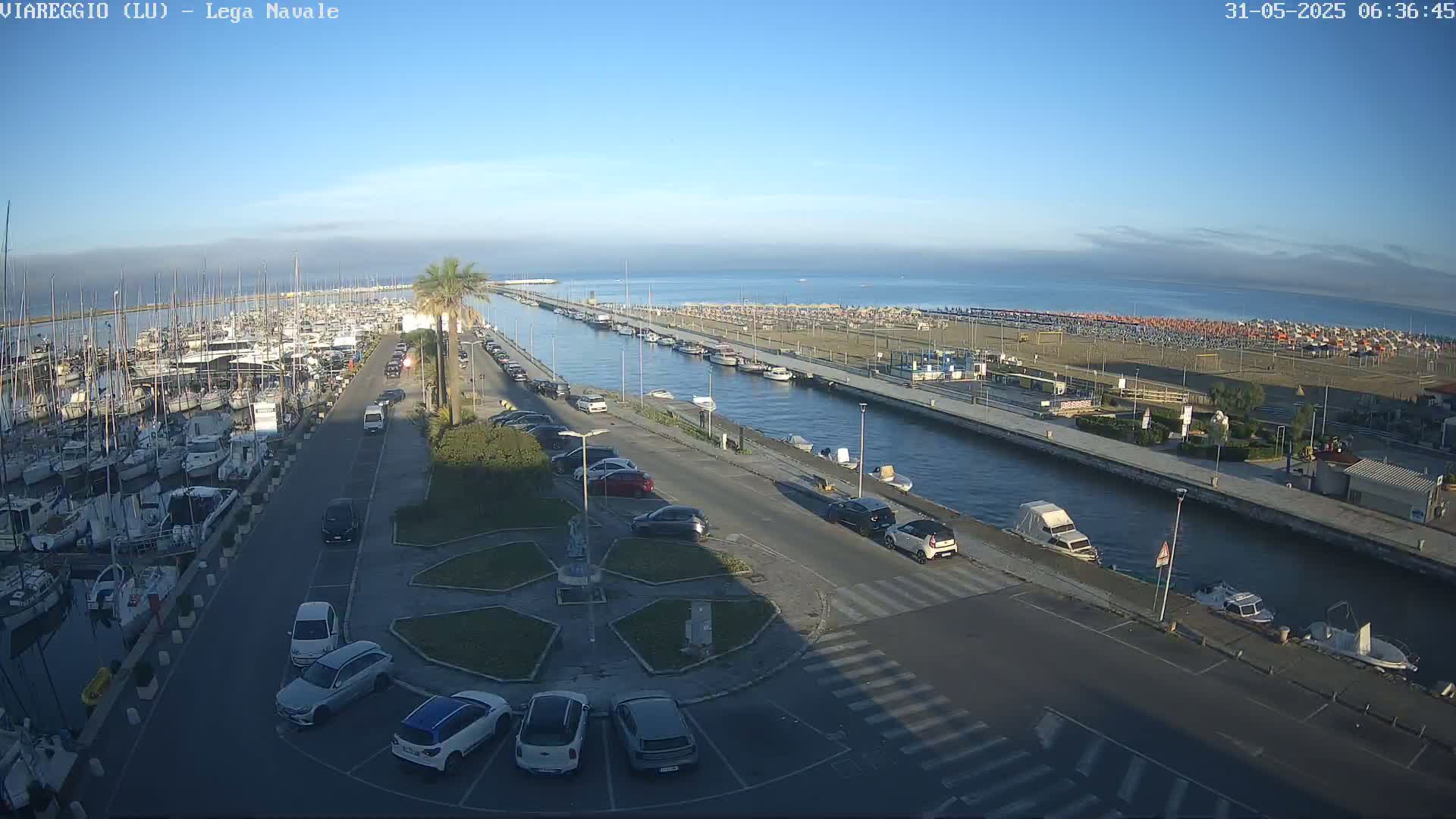 A marina filled with boats, a paved area with parked cars, and a sandy beach with numerous umbrellas are visible under a clear, sunny sky.