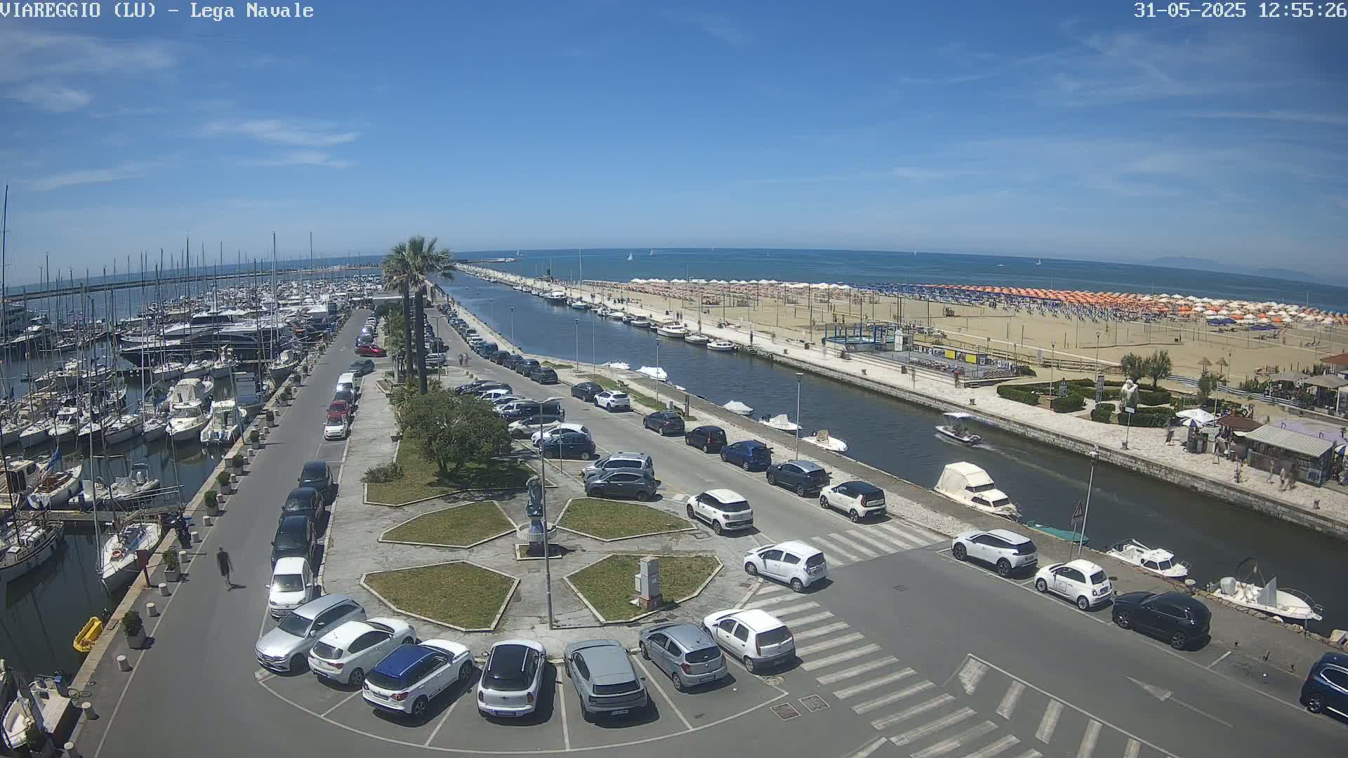 A marina filled with boats is next to a canal that leads to a sandy beach with numerous umbrellas under a clear blue sky.
