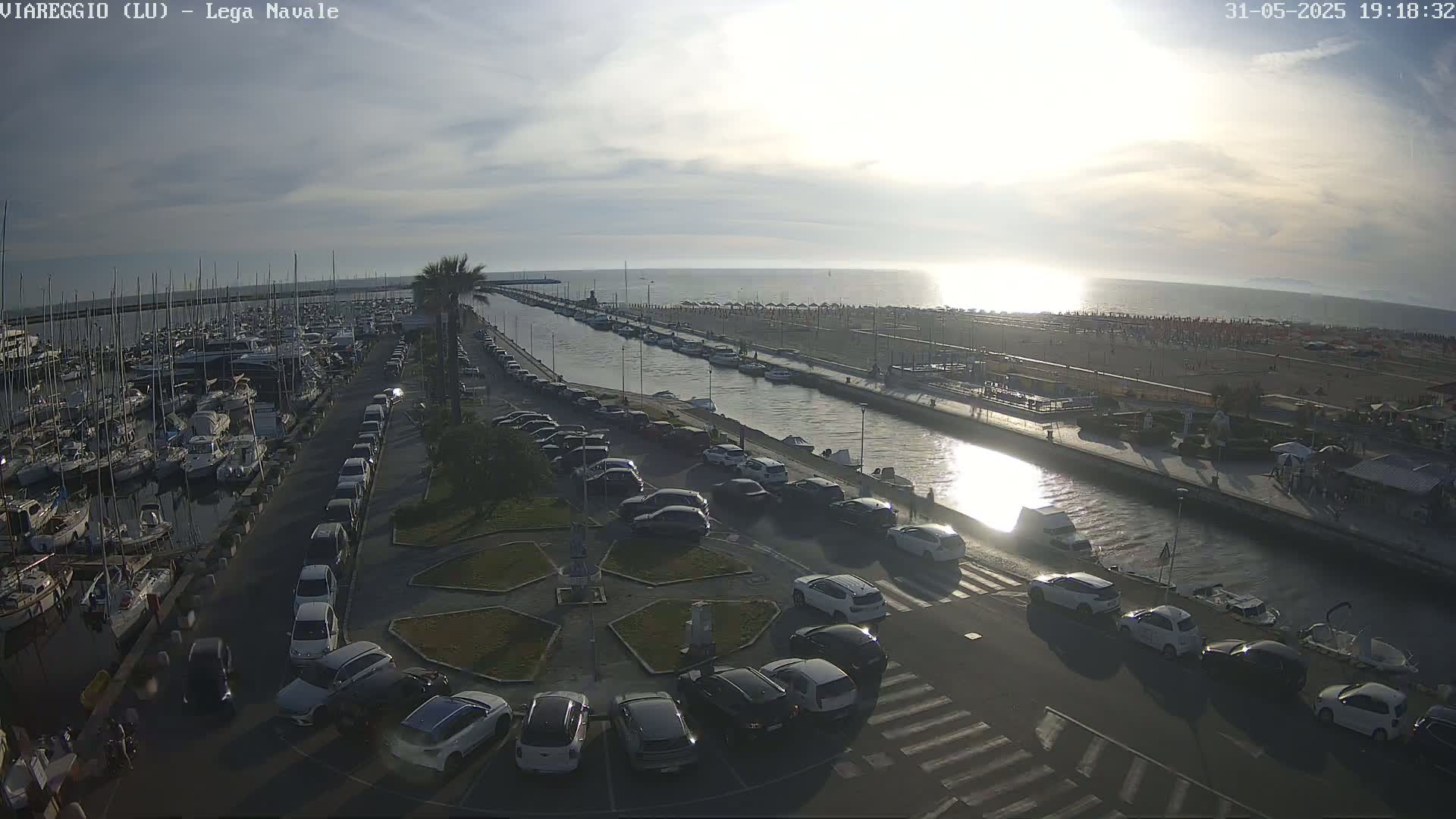 An aerial view of a marina filled with boats, a canal, a beach with people, and a parking lot filled with cars under a partly sunny sky.