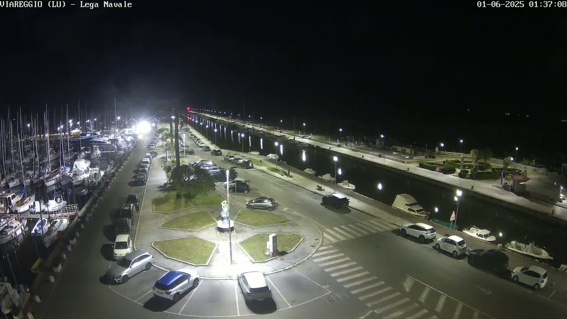 A nighttime aerial view of a marina with many boats docked, a parking lot with several cars, and a small roundabout in the center.