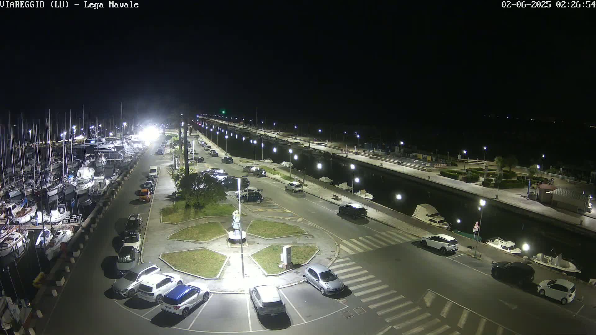 A nighttime aerial view of a marina with numerous boats docked, a parking lot filled with cars, and a circular plaza in the center, all illuminated by artificial lights.